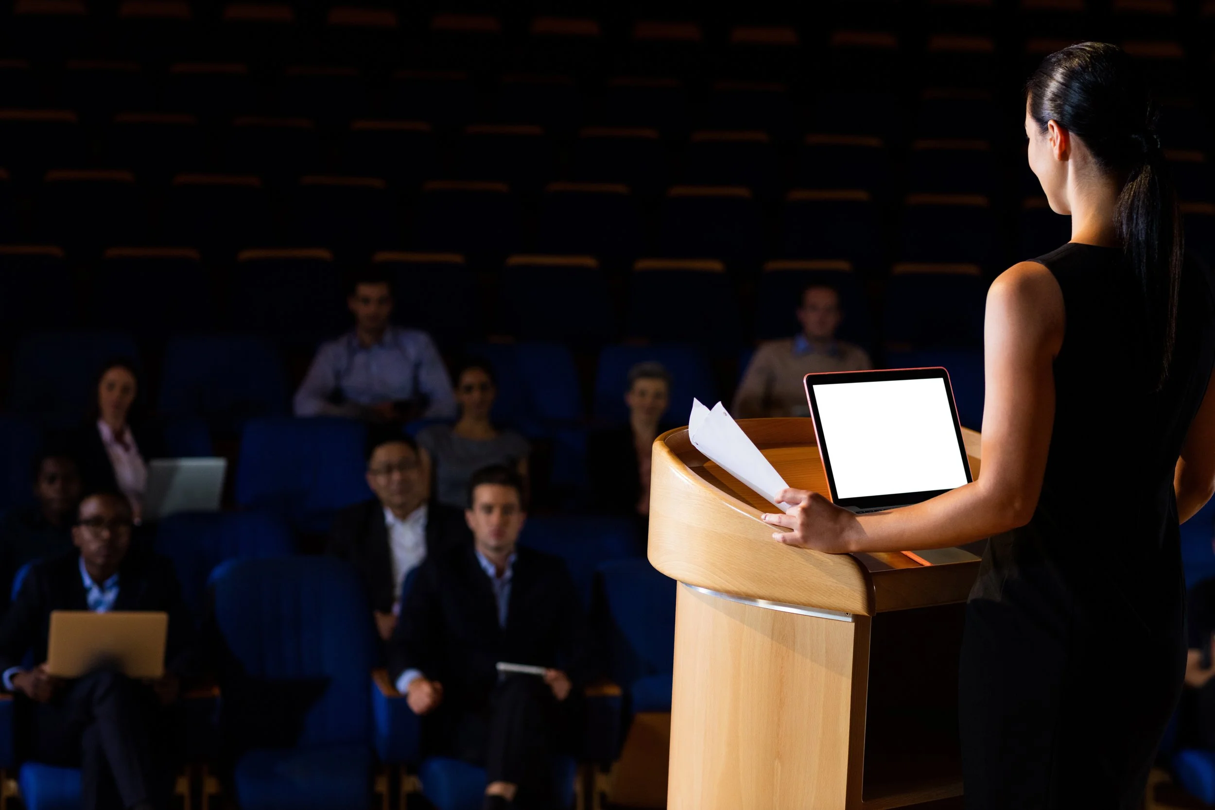 A woman in black dress giving a presentation in front of an audience in a dimly lit auditorium, standing at a wooden podium with a tablet and papers.