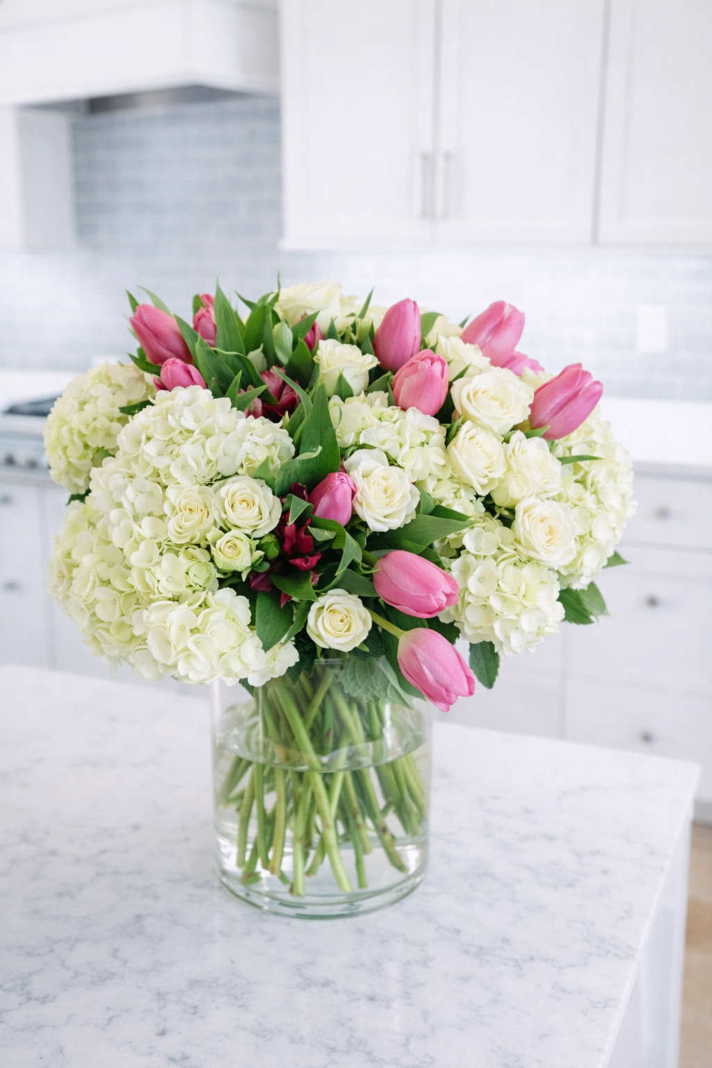 A bouquet of white roses, pink tulips, and white hydrangeas in a glass vase on a white marble countertop in a modern kitchen.