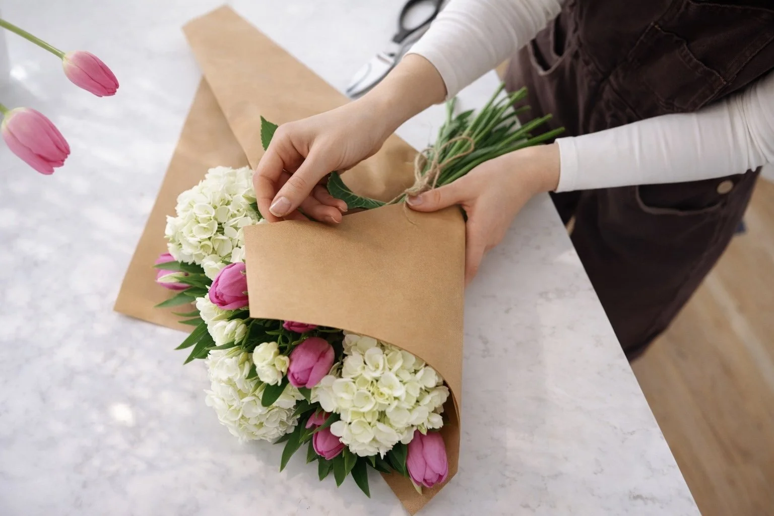 Person wrapping a bouquet of pink tulips and white hydrangeas in brown paper on a white marble surface.