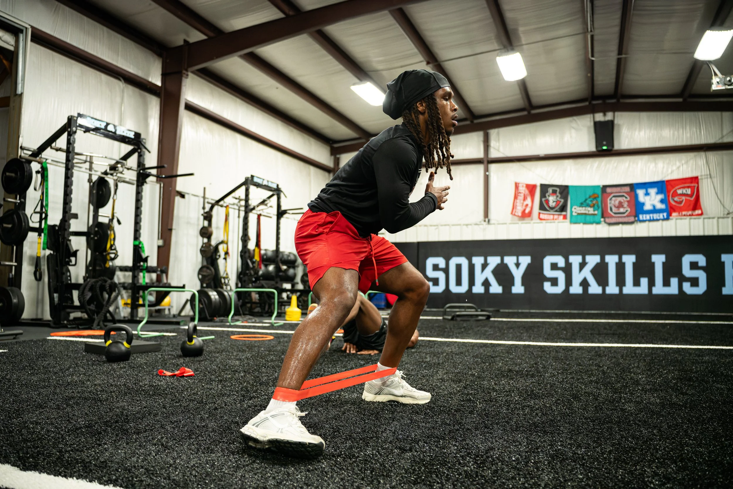 A young man in workout attire performing an exercise with resistance bands in a gym. The gym has workout equipment, blackout knee stands, and banners hanging on the wall in the background.