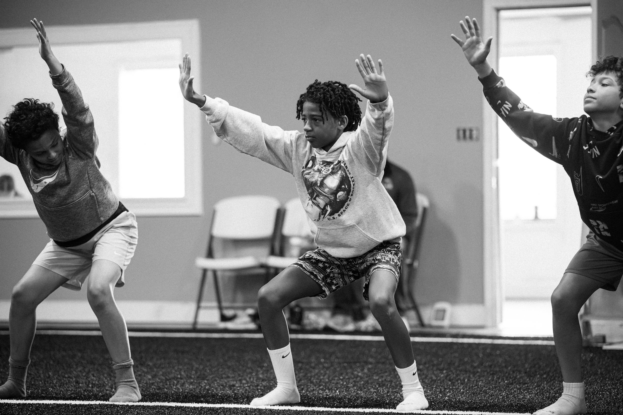 Three young boys practicing dance moves in a room with chairs and windows in the background.