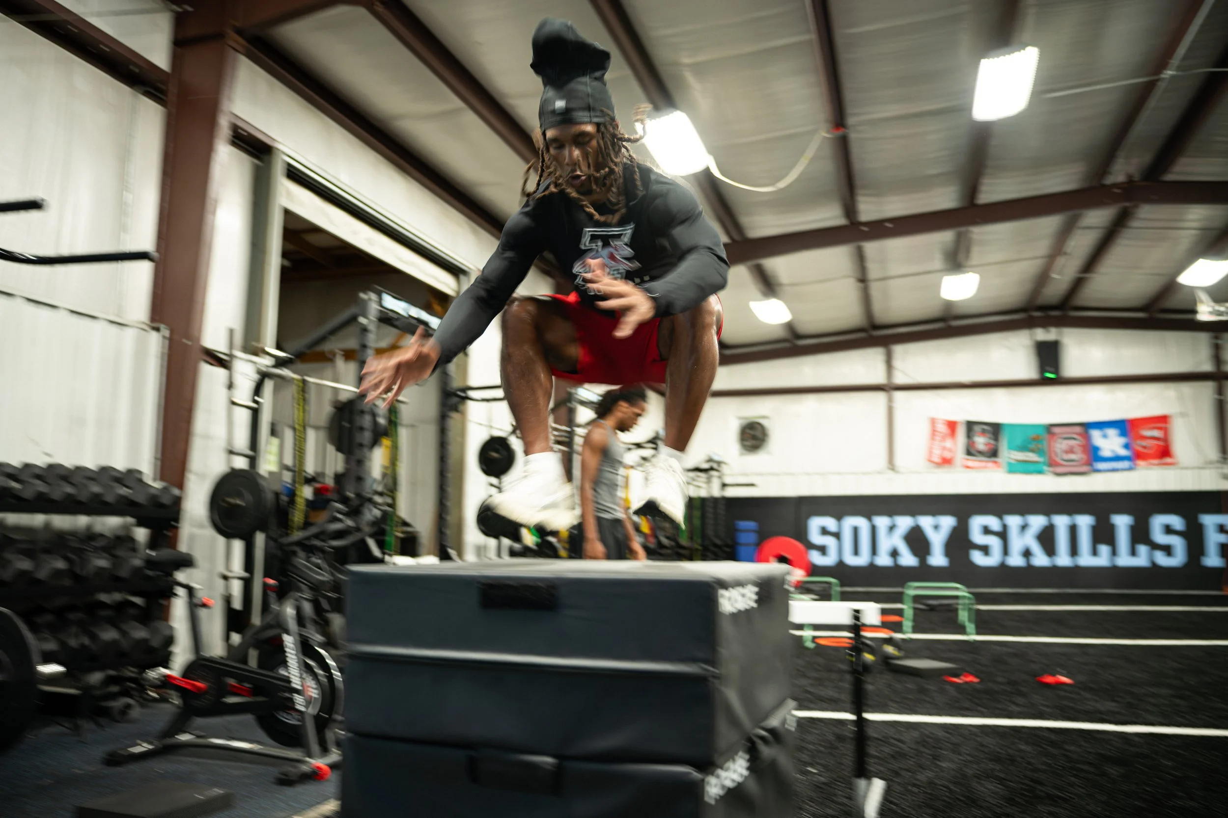 A person with dreadlocks wearing a black hoodie and red shorts jumping in an indoor gym, with gym equipment and another person in the background.