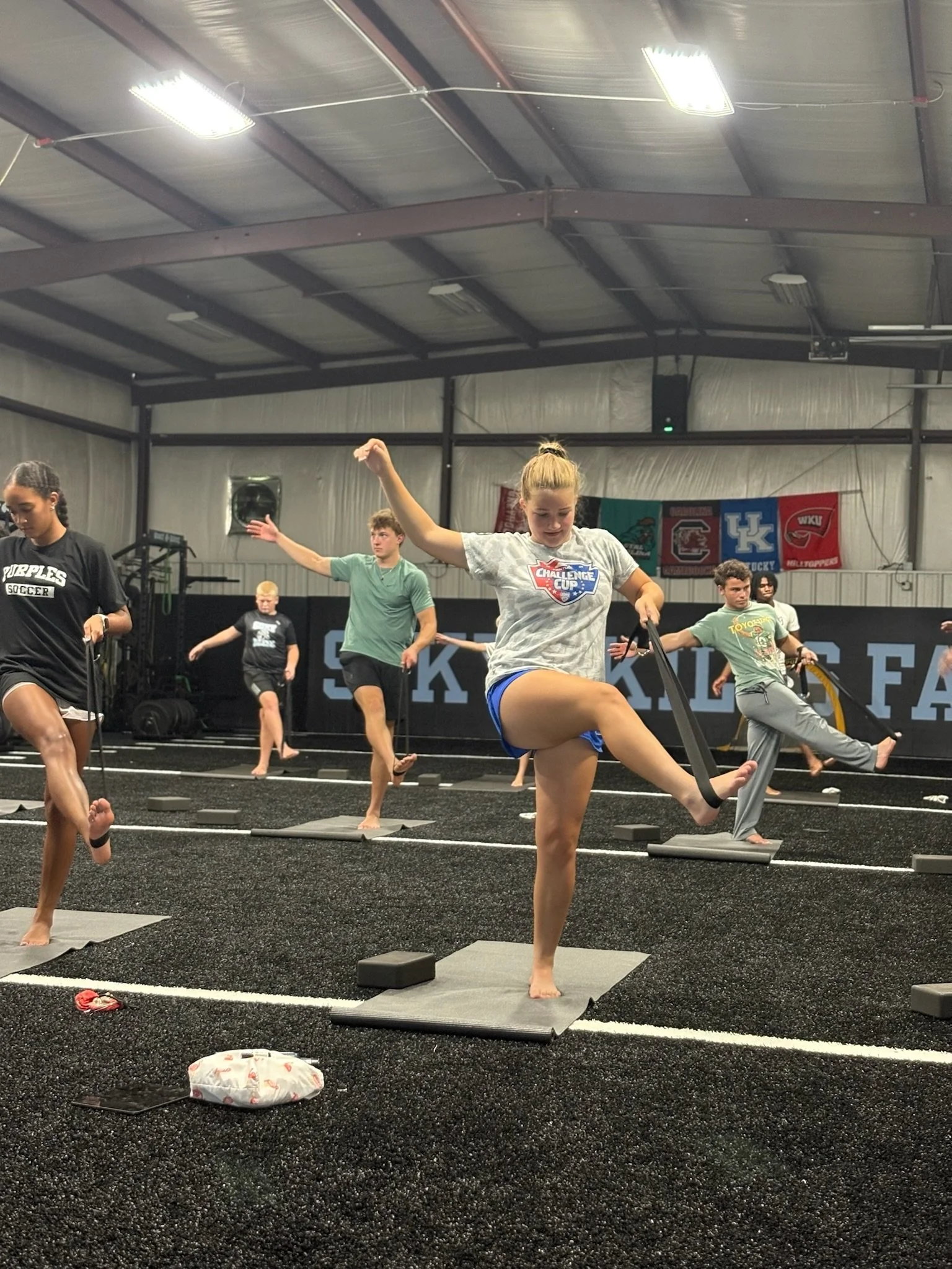Young women and men participating in a fitness class indoors, performing balancing exercises on mats, some with resistance bands, the environment appears to be a gym with banners and equipment in the background.