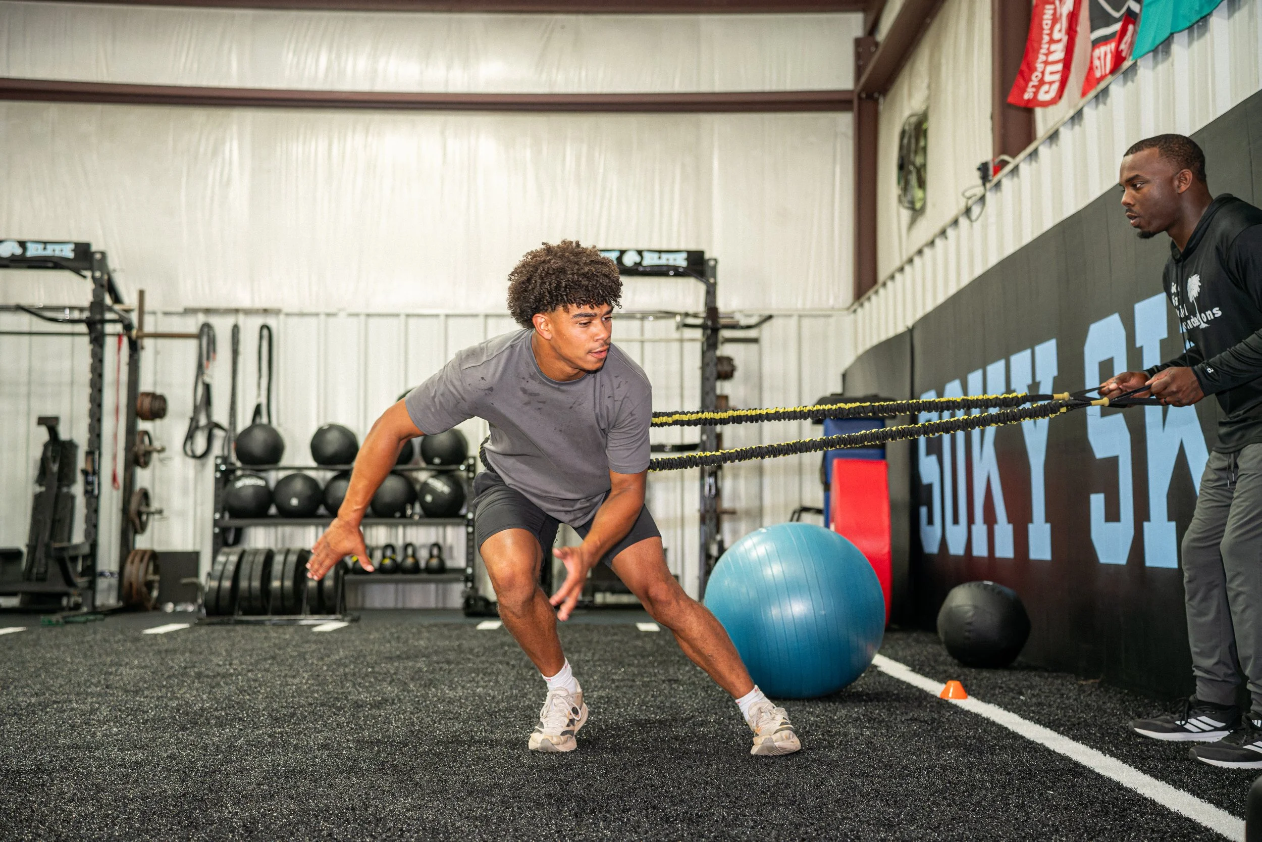 Two men working out in a gym. One man is performing an exercise using a resistance band operated by the other man, with various gym equipment in the background.