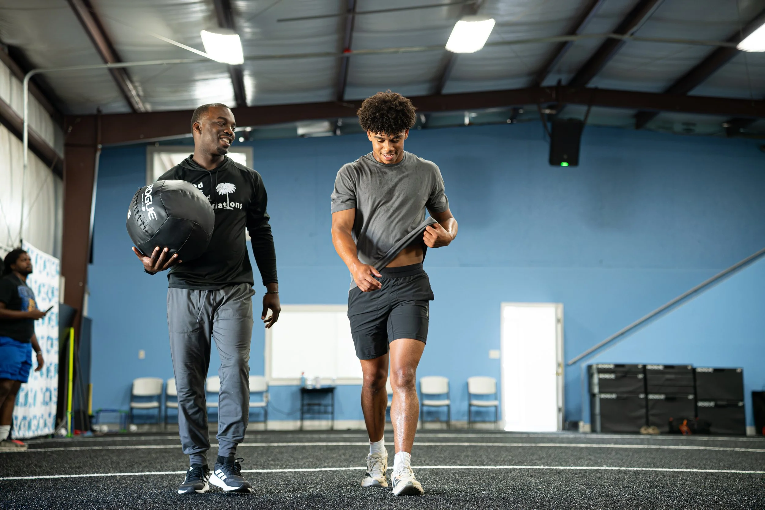 An athletic young man in a gray t-shirt and black shorts walking on a track inside a gym, with a coach holding a medicine ball and smiling beside him.