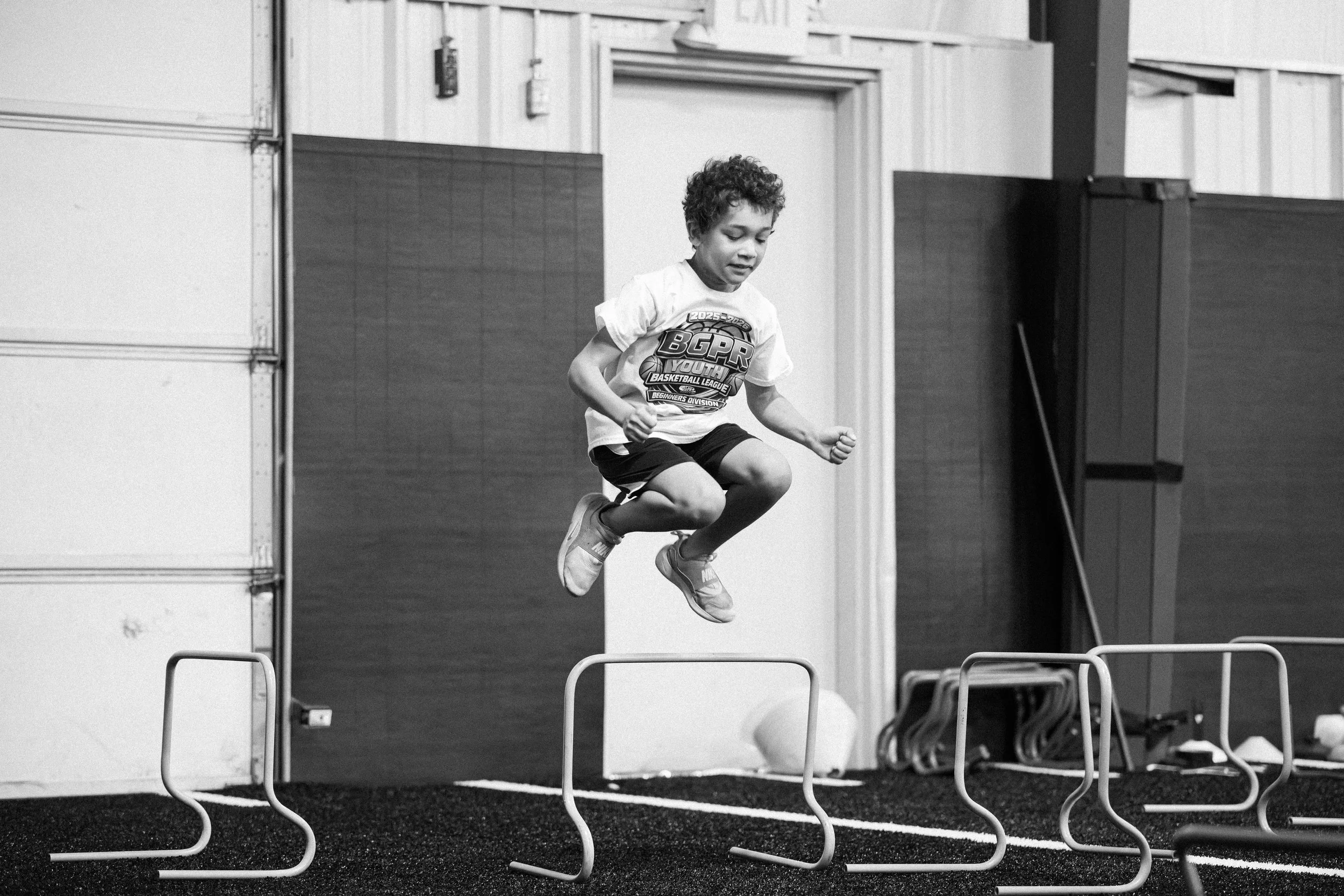 A boy with curly hair jumping over hurdles during a training session or sports event, in an indoor setting.