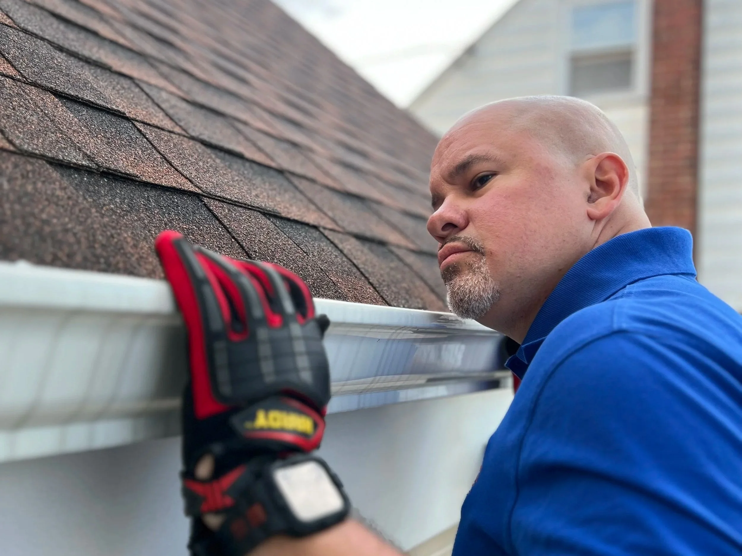 James Morales from Arc light home inspections performing a residential home inspection inspecting a gutter on a house roof. He is wearing a black and red glove on his left hand and has a focused expression.