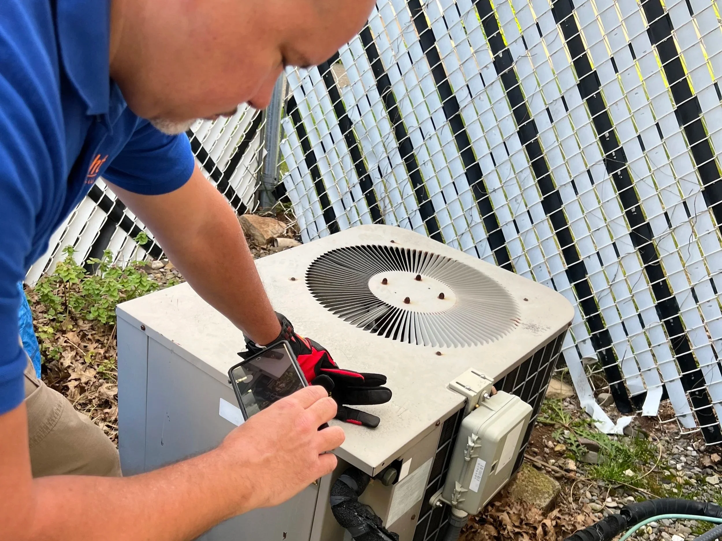 James Morales from Arc light home inspections performing a residential is taking a photo of an outdoor air conditioning unit with a smartphone. The unit is on the ground, surrounded by leaves, with a white and black lattice fence in the background.