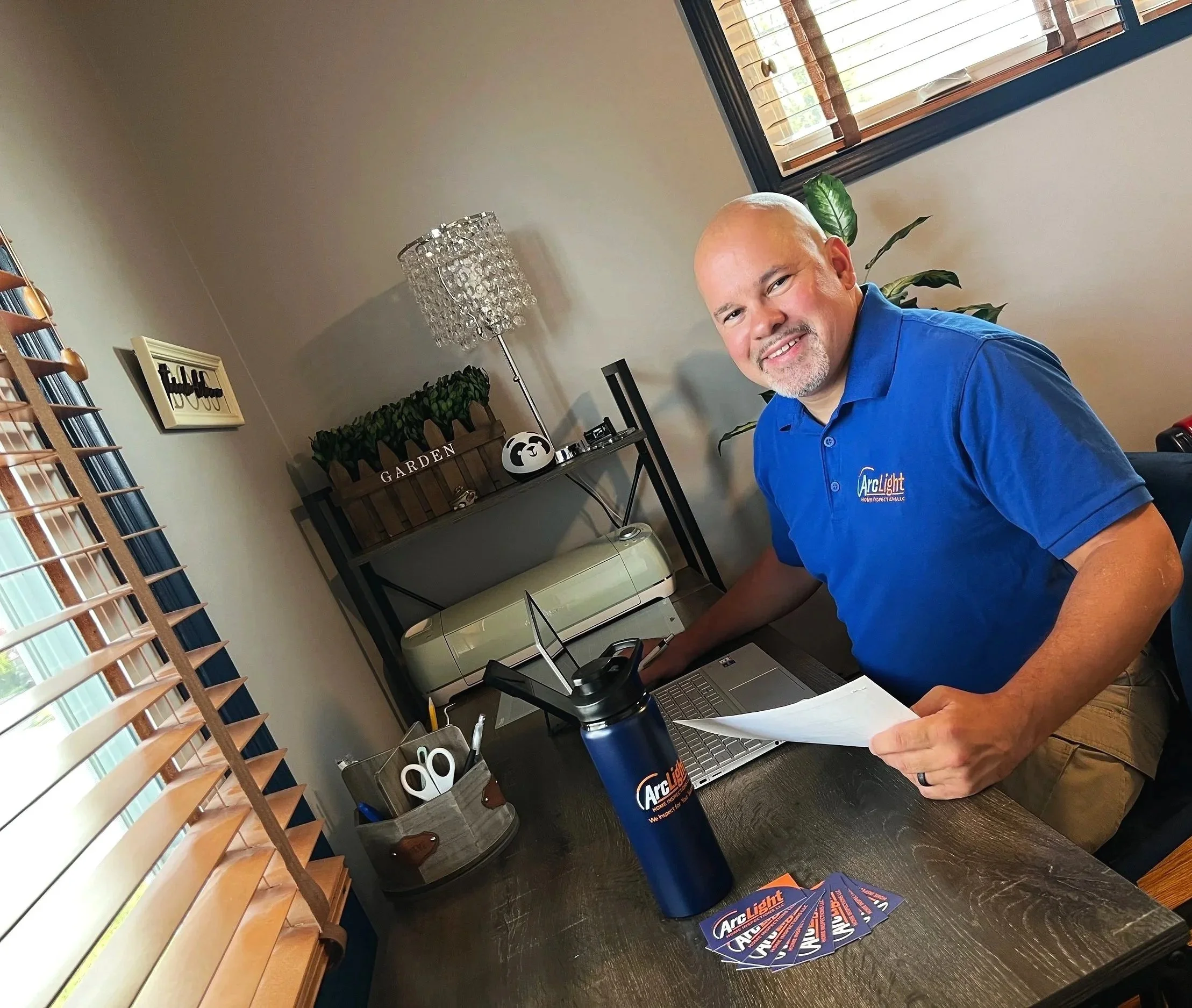 James Morales Arc Light Home Inspections The desk has a laptop, a blue water bottle, and business cards. He is smiling and holding a printed document.