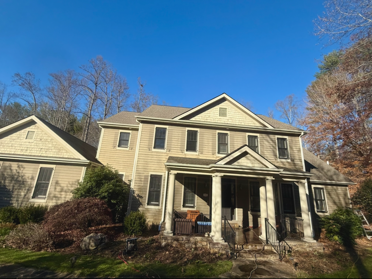 A beige, multi-story house with a porch supported by white columns, set against a clear blue sky and surrounded by trees, some with fall foliage.