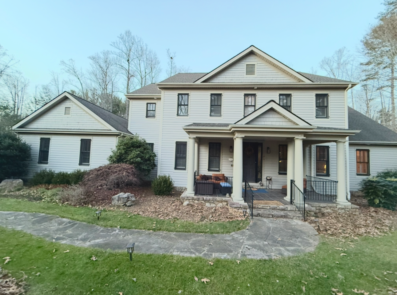 A large two-story white house with black shutters and a porch with pillars, surrounded by trees and a landscaped yard with bushes and rocks.
