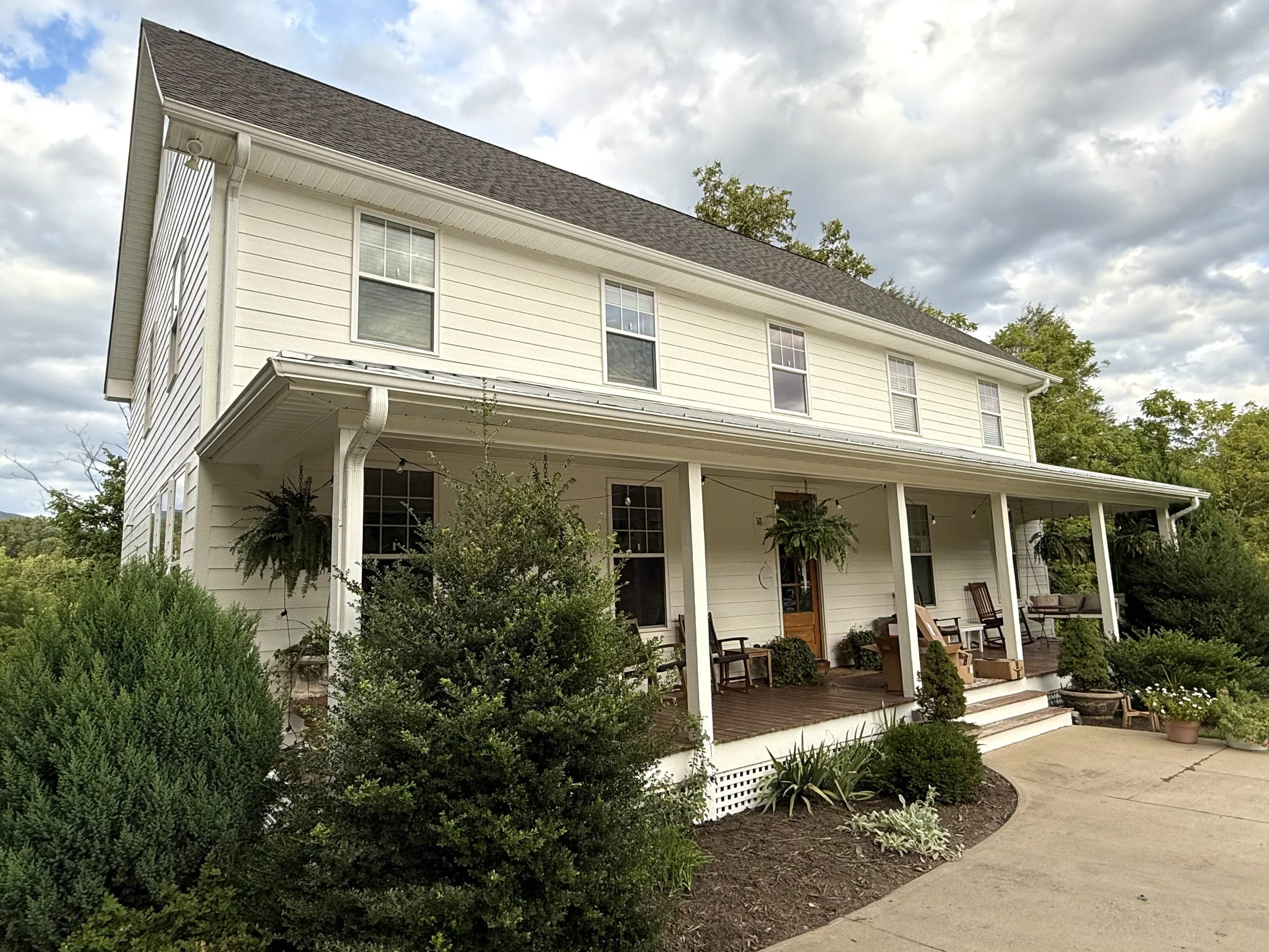 Front view of a two-story white house with a porch, surrounded by greenery and potted plants, under a partly cloudy sky.