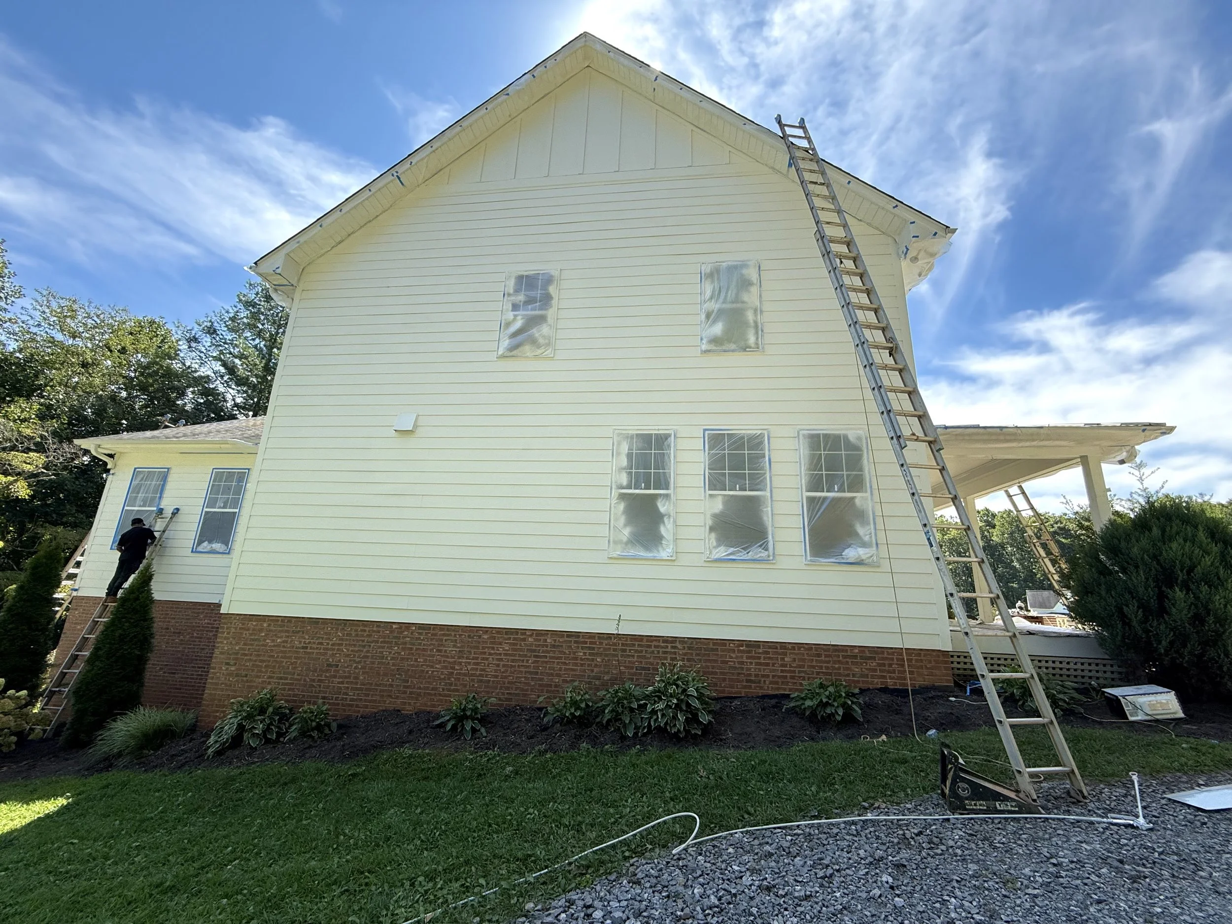 Exterior of a house with yellow siding and a brick foundation, being painted white, with two workers painting the upper walls, ladders for access, and a mostly clear blue sky with some clouds.