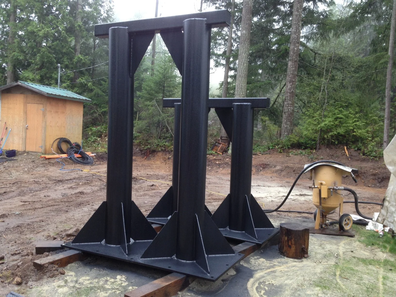 Large black metal structure with three vertical supports on a construction site with trees and a small shed in the background.