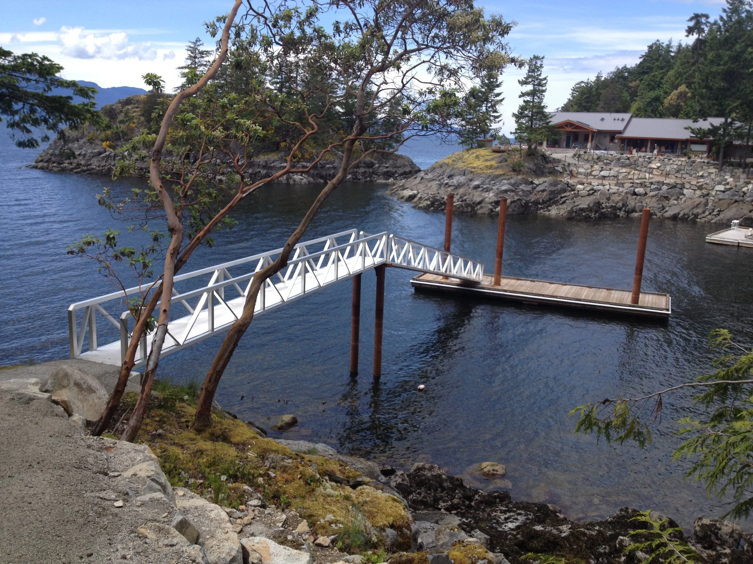 A small wooden pier with a white metal bridge leads over water, with a rocky shoreline and trees, and a building on a hill in the background.