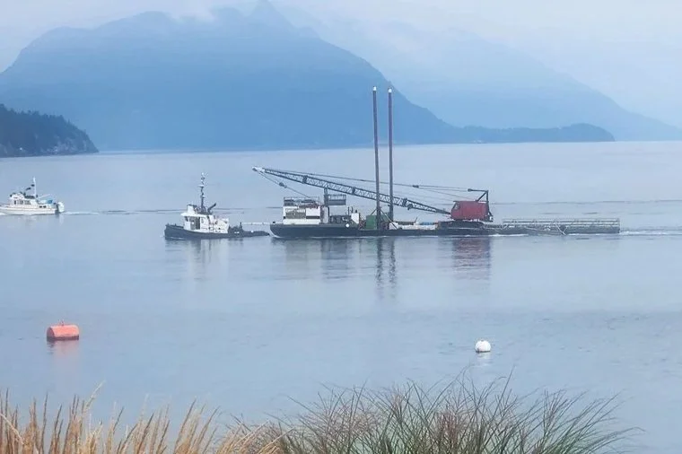 A barge on a calm lake with a crane and small boats, surrounded by mountains in the background.