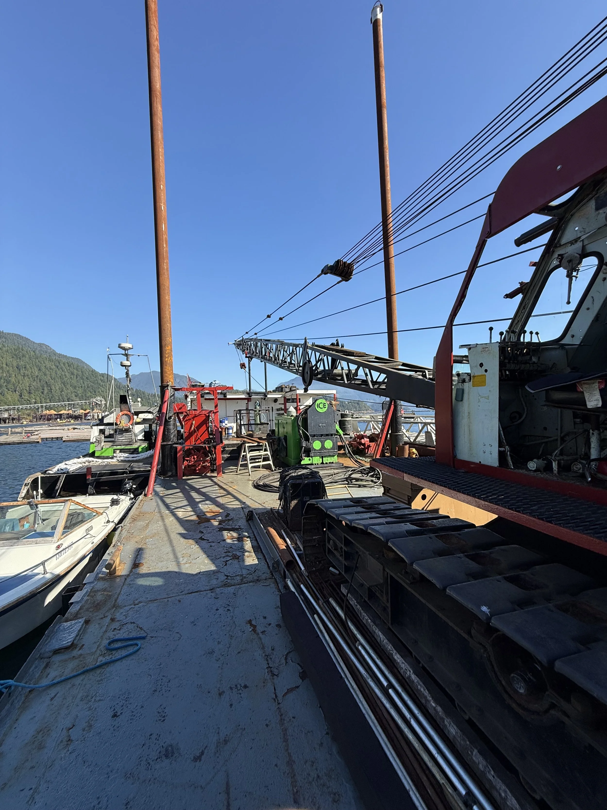 View of a boat deck with equipment, hoses, and machinery, with a crane, and a marina with boats and water in the background under a clear blue sky.
