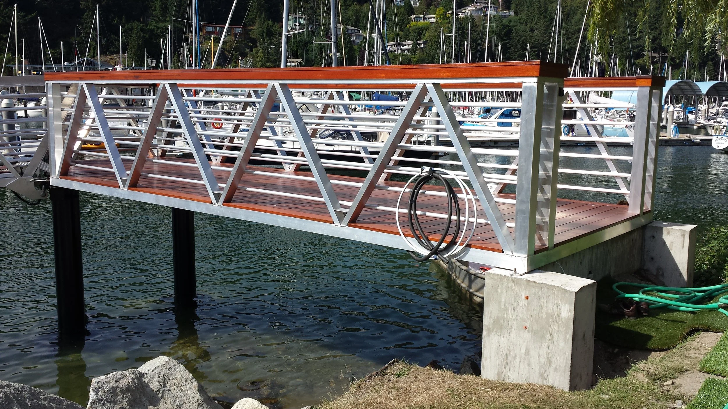 A metal and wood dock extending over water at a marina, with boats and sailboats moored in the background.