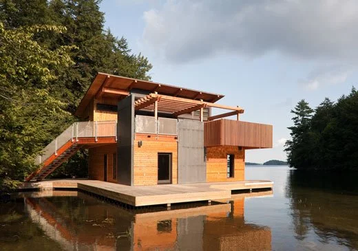 Modern house on the water with wooden and concrete exterior, large windows, and a deck, surrounded by trees.