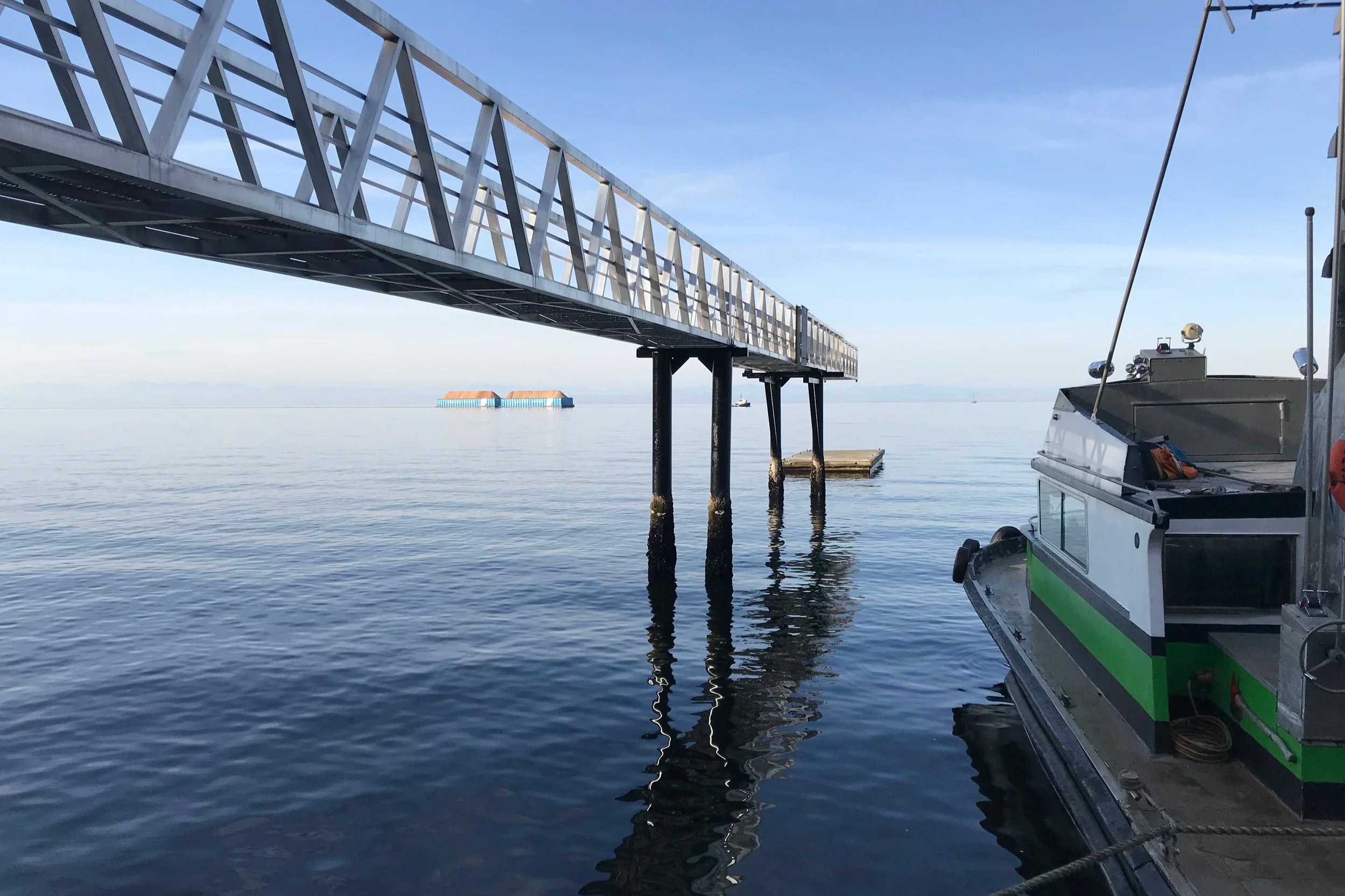 A metal pier extending over calm water with a boat docked to the right side. In the background, there is a floating platform with orange rooftops and a small boat nearby, under a clear blue sky.