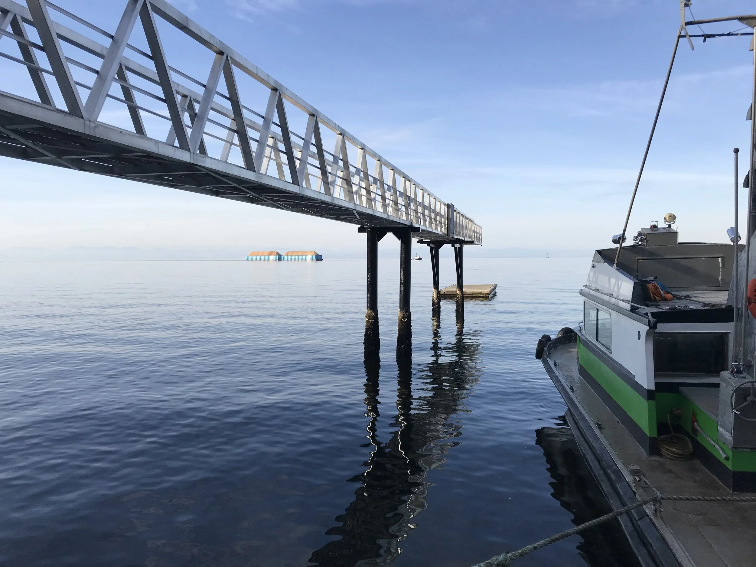 A dock extending into calm water with a boat moored on the right side and a floating platform in the distance under a clear blue sky.