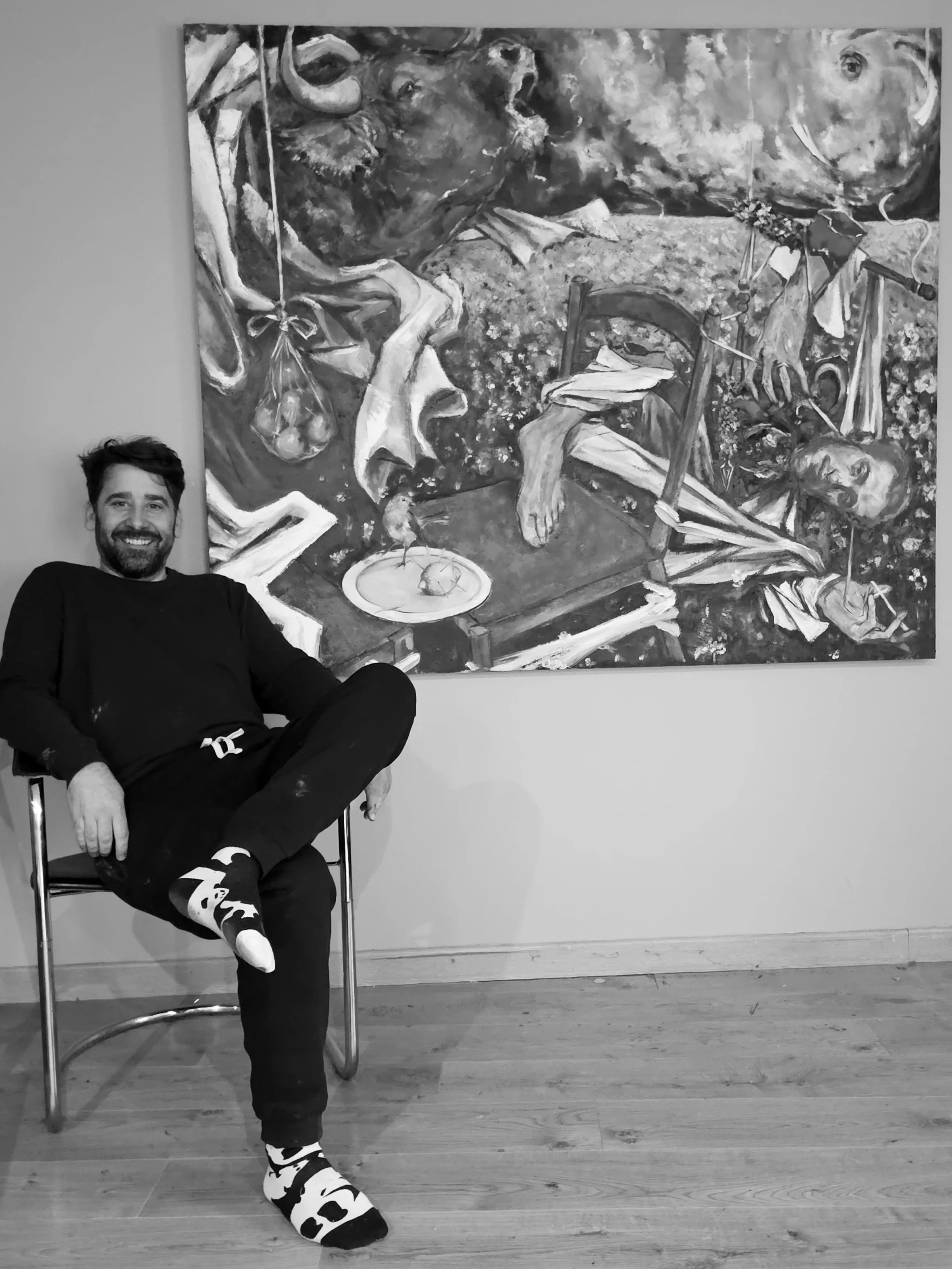 A man sitting on a chair with one leg crossed over the other, smiling at the camera, in front of a large abstract painting on a wall.