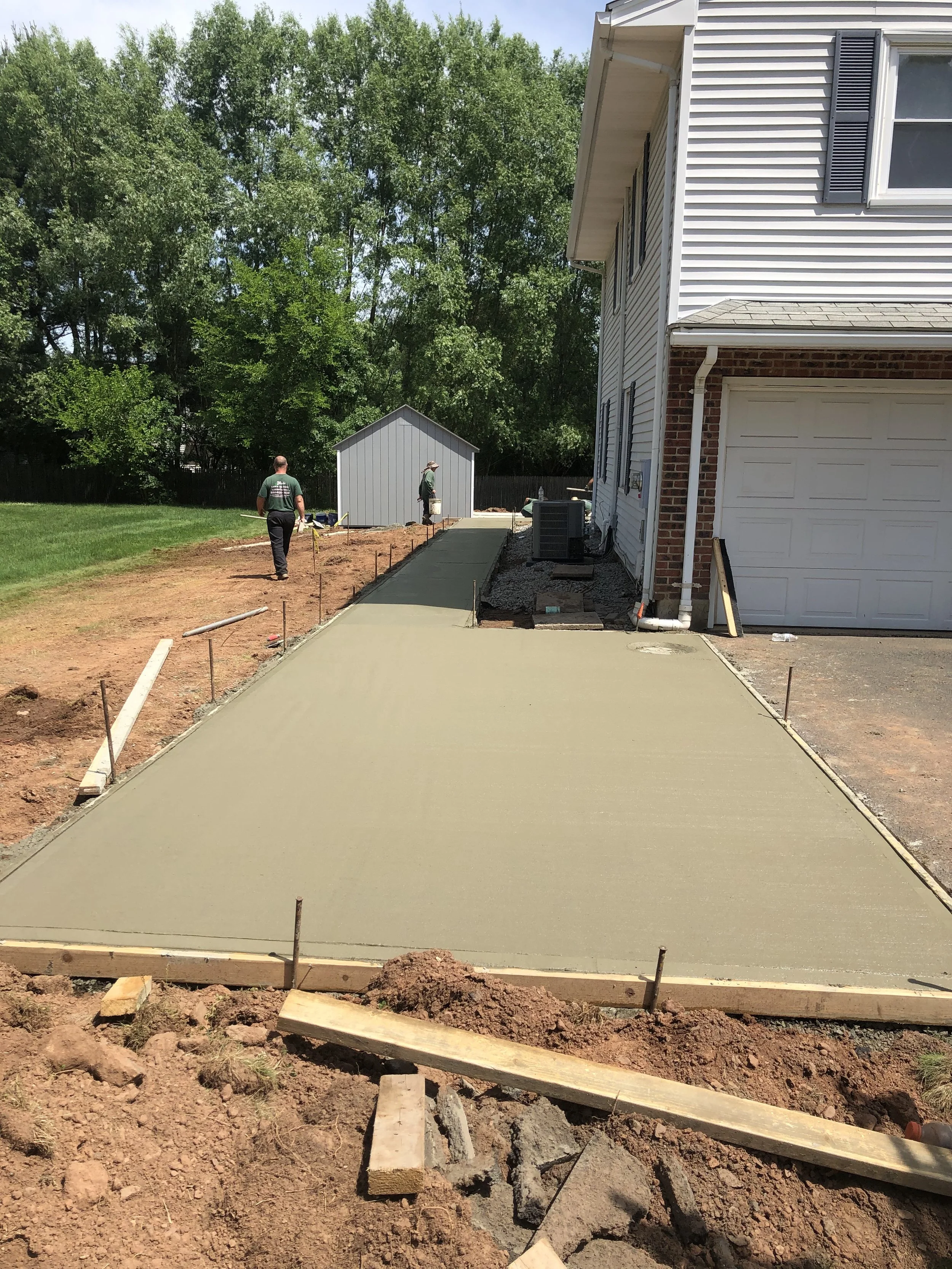 Two workers are pouring new concrete for a sidewalk in a residential backyard, with a house on the right and a small shed in the background, surrounded by a tree-lined yard.