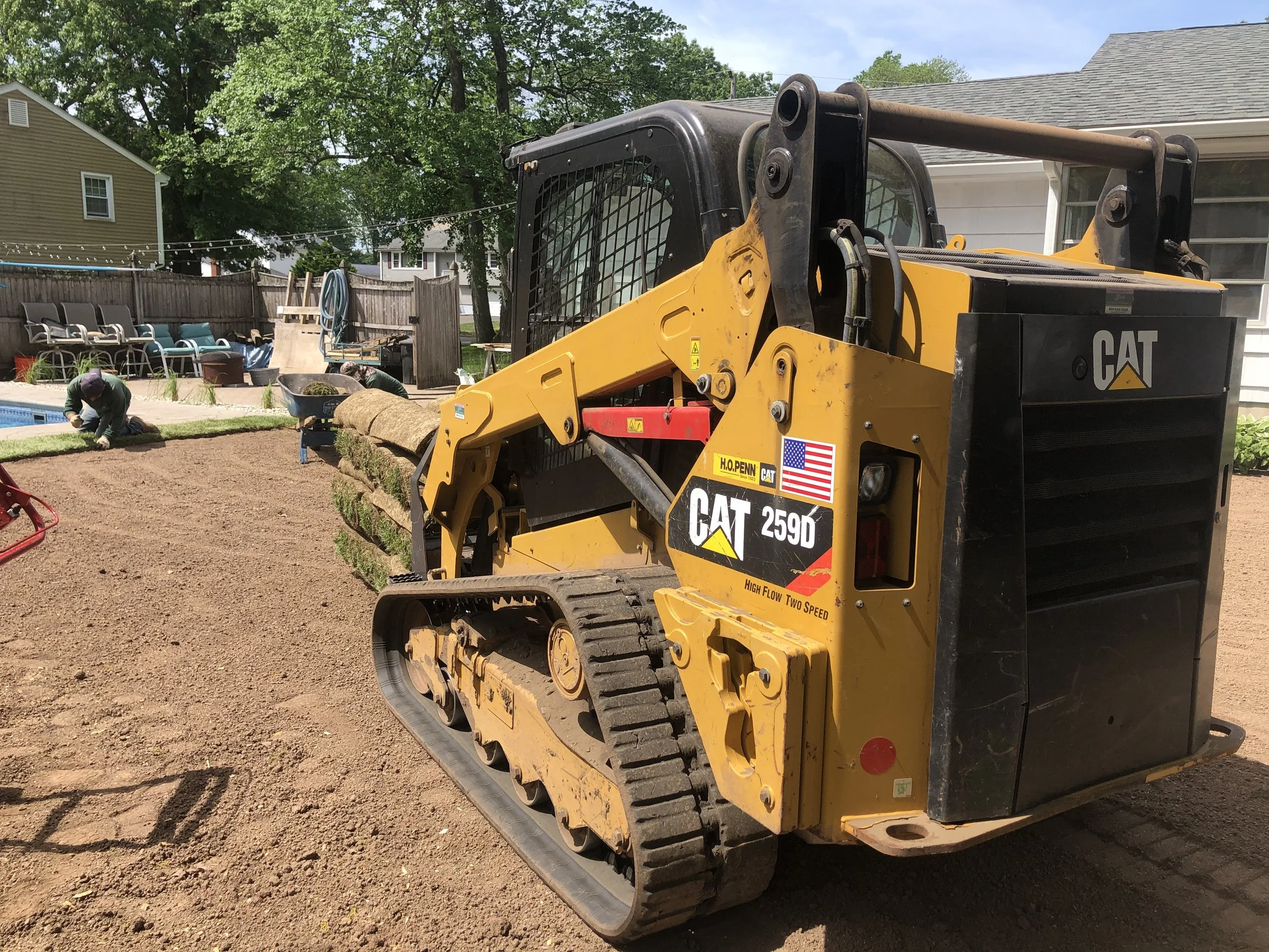 A yellow CAT 259D compact track loader working on landscaping in a backyard, with workers spreading soil and assembling a garden area.