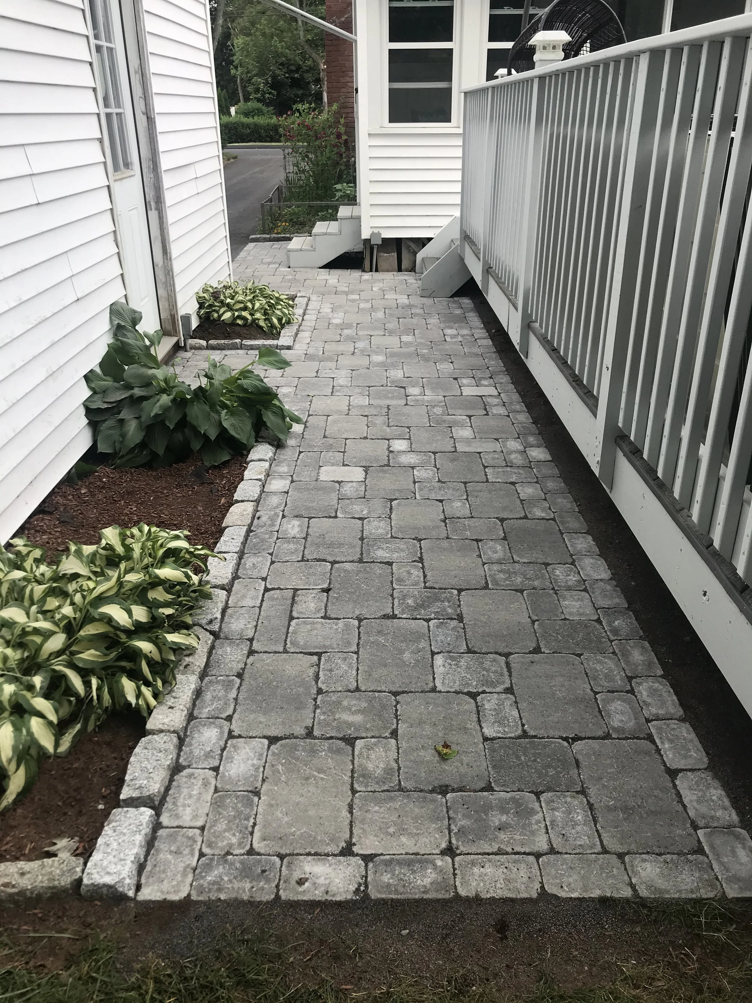 A paved walkway made of interlocking gray bricks, bordered by flower beds and a white railing on the right side, leading to the steps of a house.