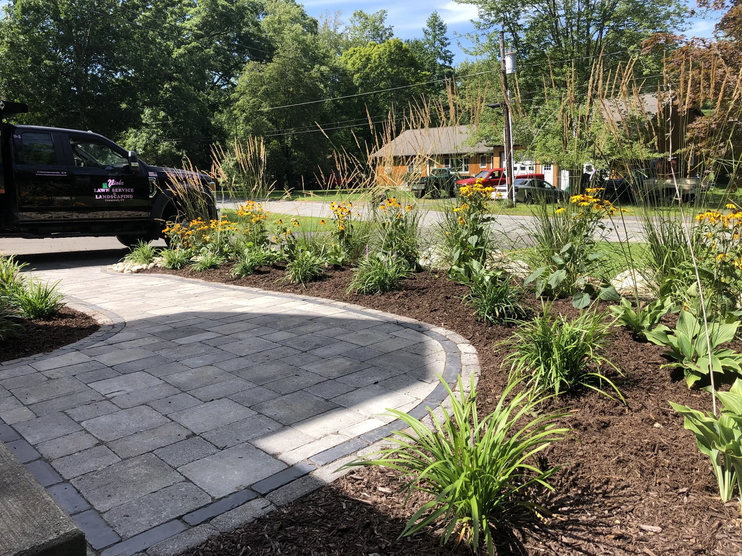 A landscaped yard with a stone pathway, various flowering plants, and a lawn area with parked cars in the background.