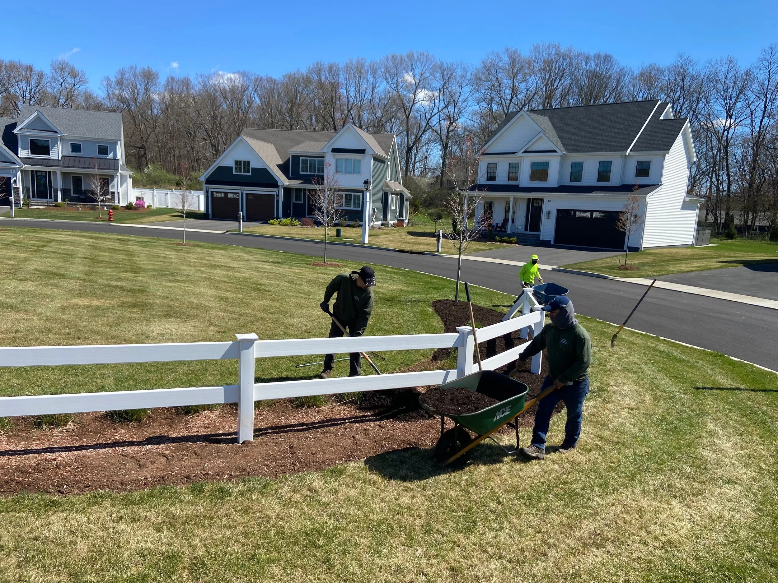 Workers planting flowers and filling a flower bed with dark soil behind a white picket fence in a neighborhood with modern houses and green lawns on a sunny day.