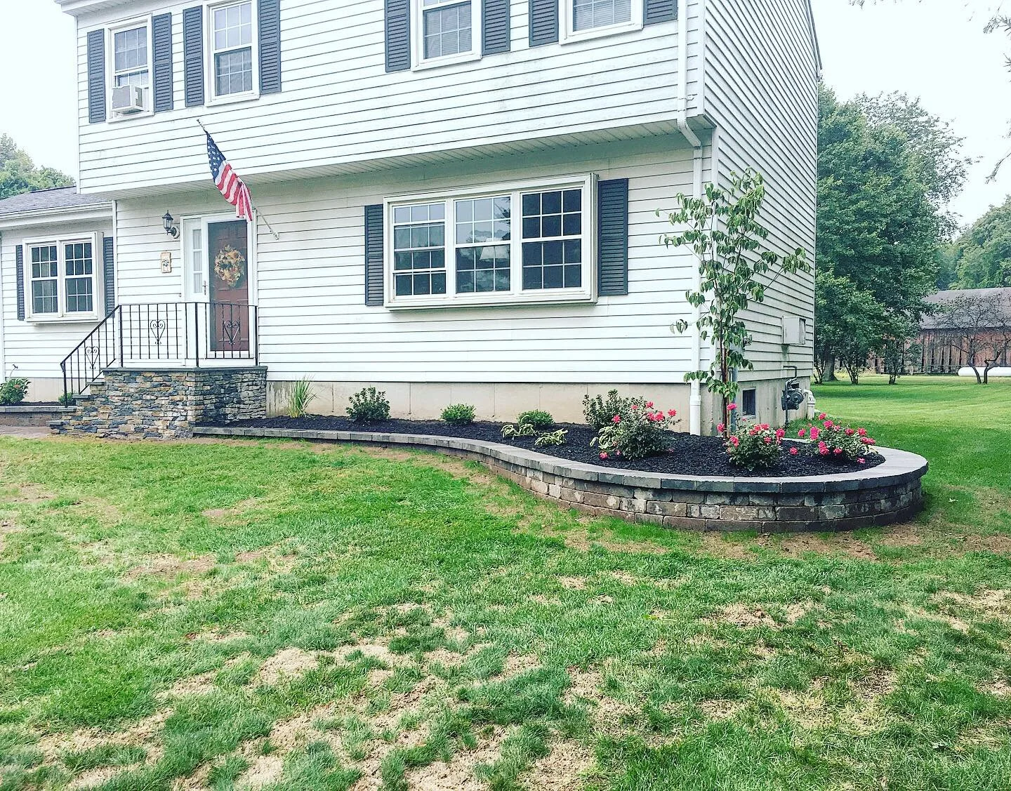 Front yard with a white house, featuring black shutters, an American flag, and a landscaped flower bed with bushes and pink flowers.