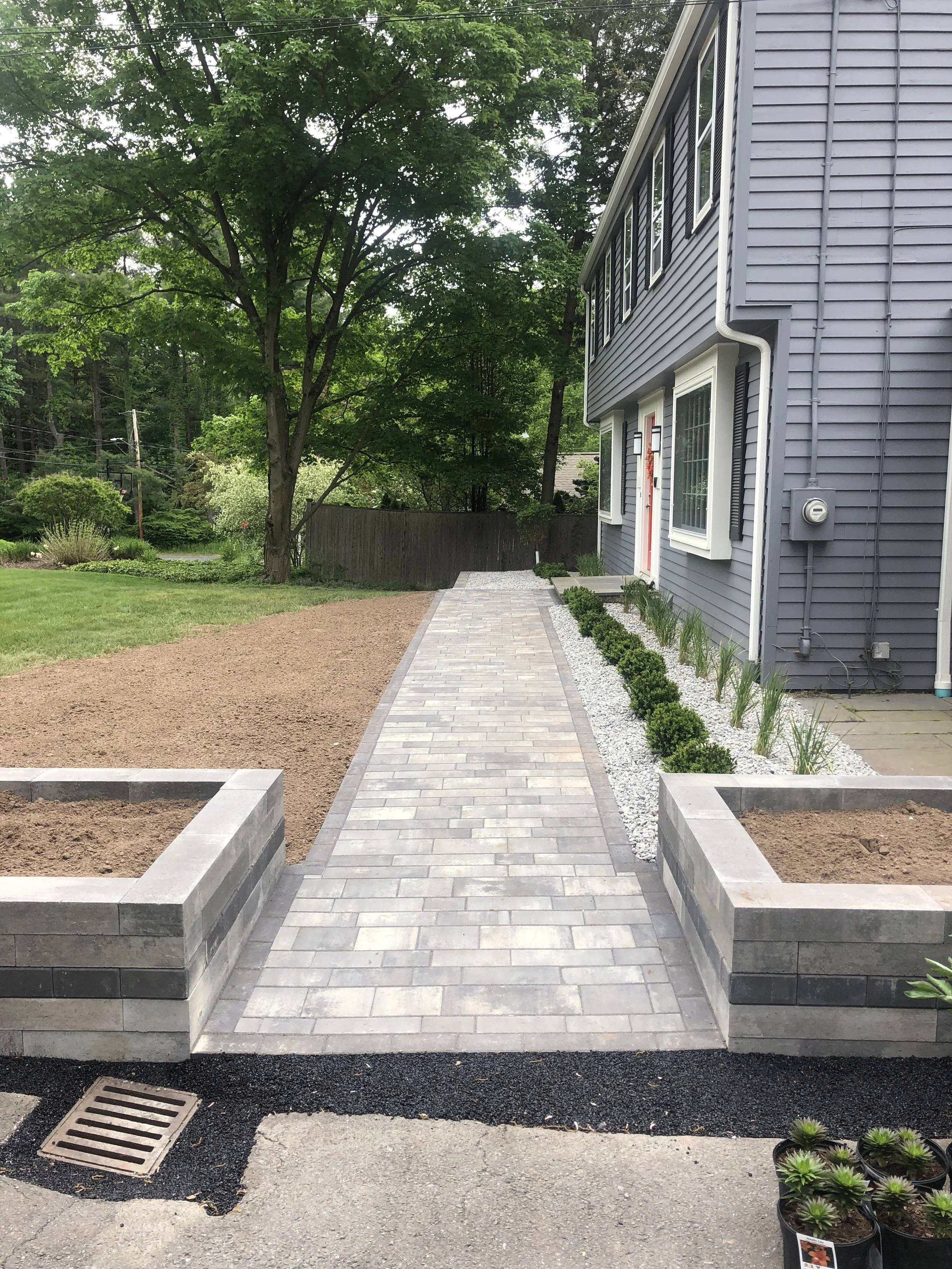 Newly paved walkway with brick pattern leading to the house, bordered by small bushes and white gravel, with a large tree and green lawn on the left and a gray house with multiple windows on the right.