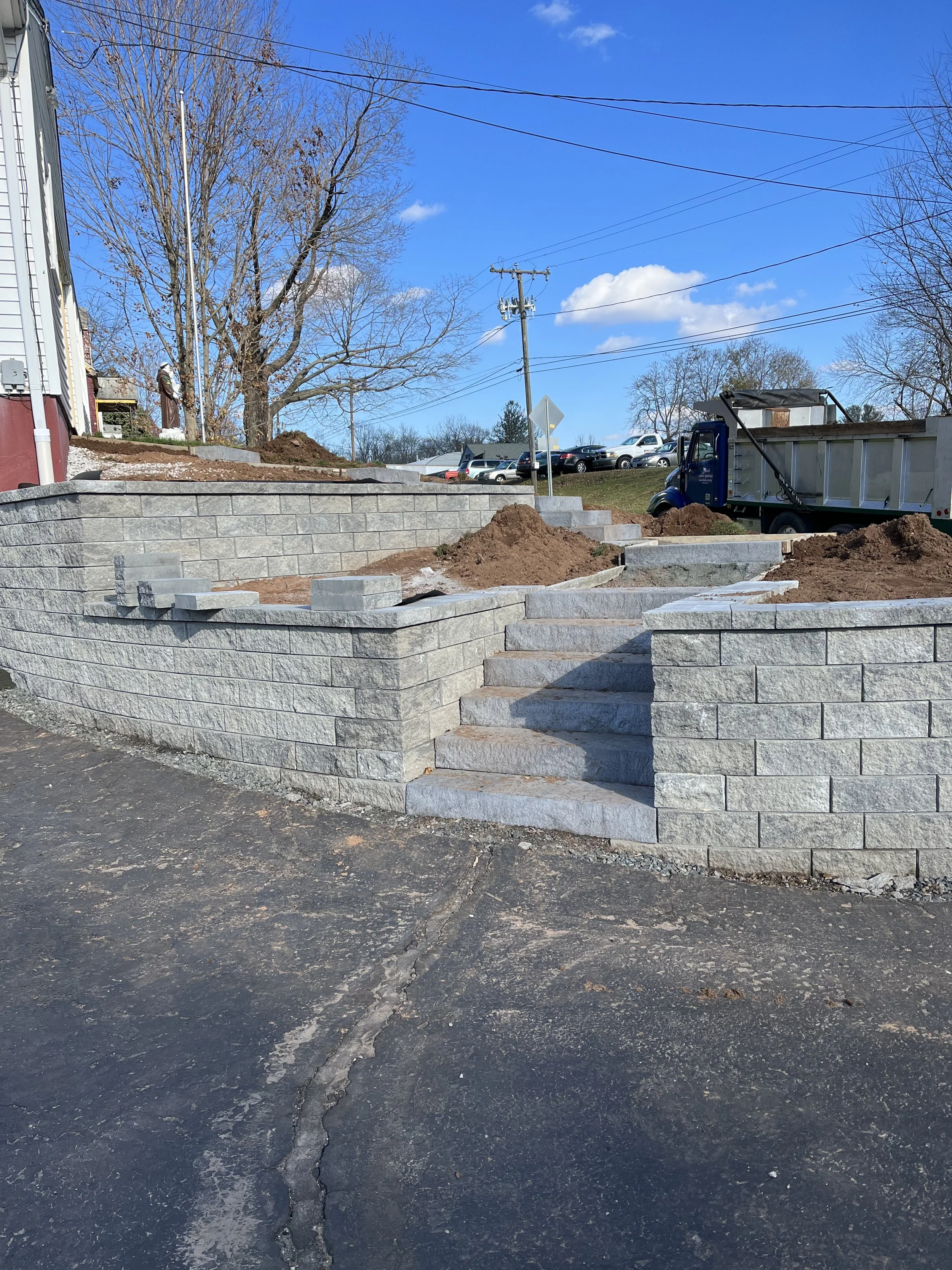 Construction site with a stone retaining wall and stairs, piles of dirt, a partly cloudy sky, leafless trees, and a parking lot with cars in the background.