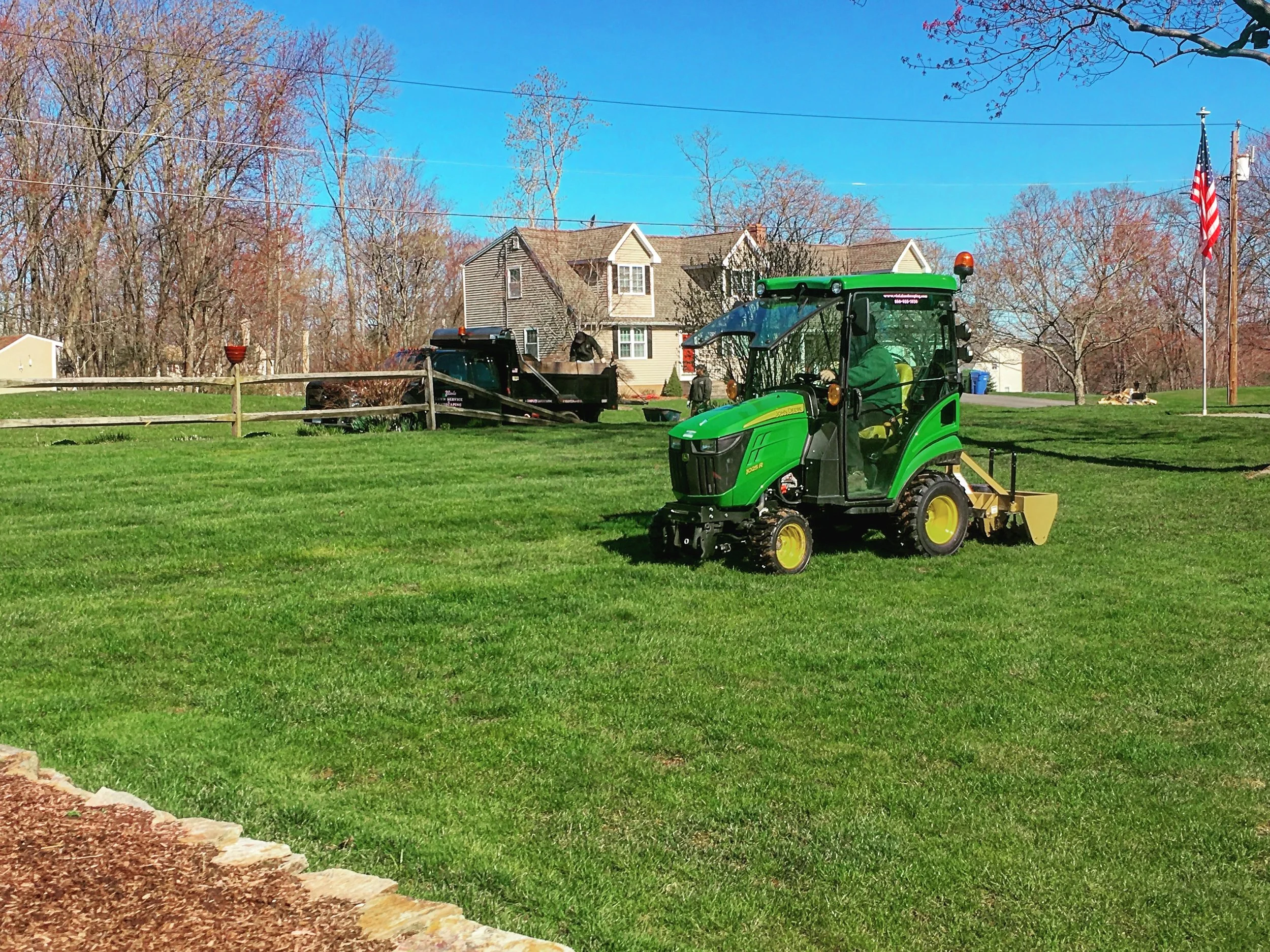 A person operating a green John Deere lawn mower on a well-maintained lawn in a suburban neighborhood with houses, trees, power lines, an American flag, and blue sky in the background.