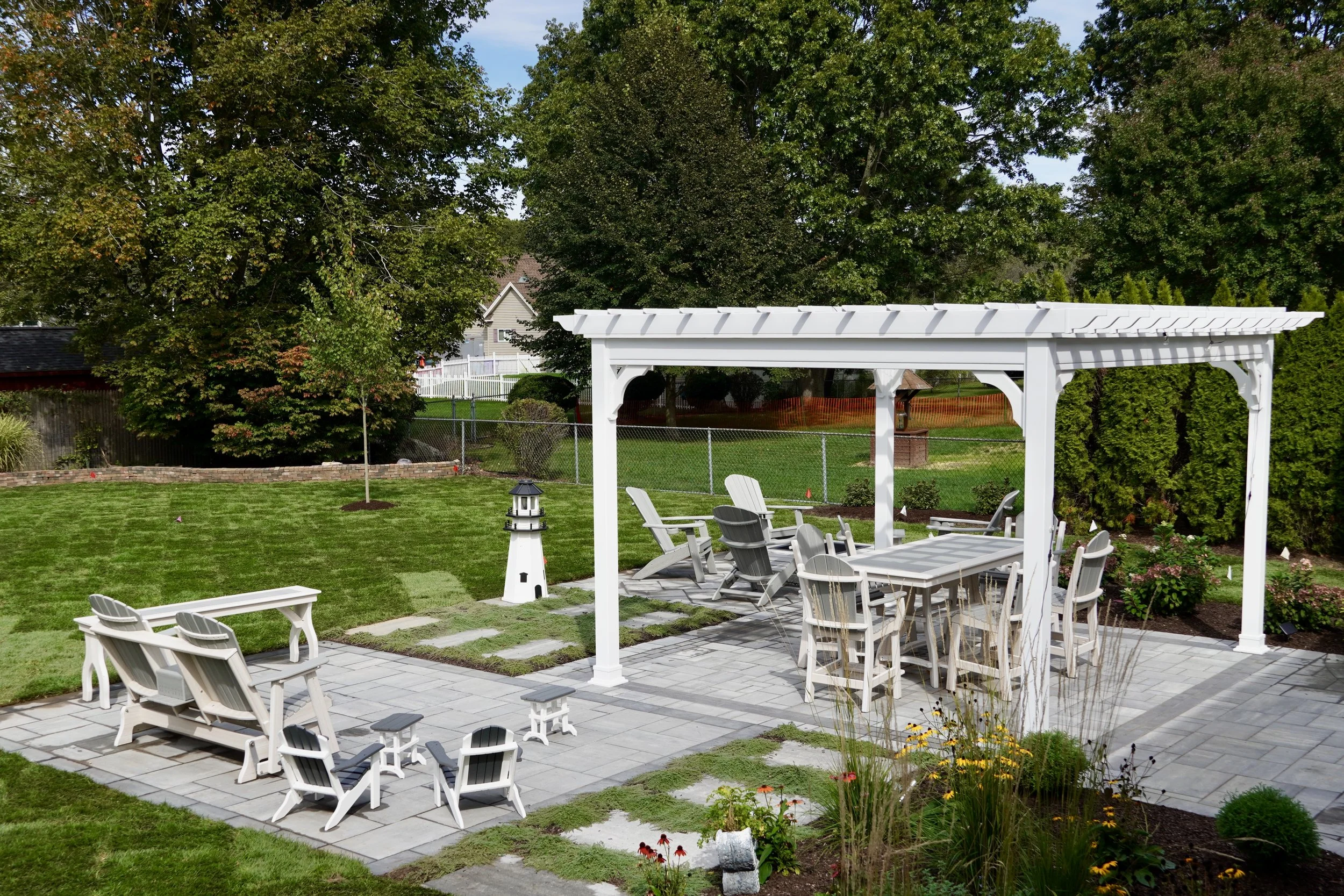 A backyard patio area with outdoor furniture, including a table with six chairs, a bench with two chairs, and Adirondack chairs, under a white pergola. There is a small lighthouse decor and a garden with flowers and shrubs. The yard has a green lawn, trees, and a chain-link fence in the background.