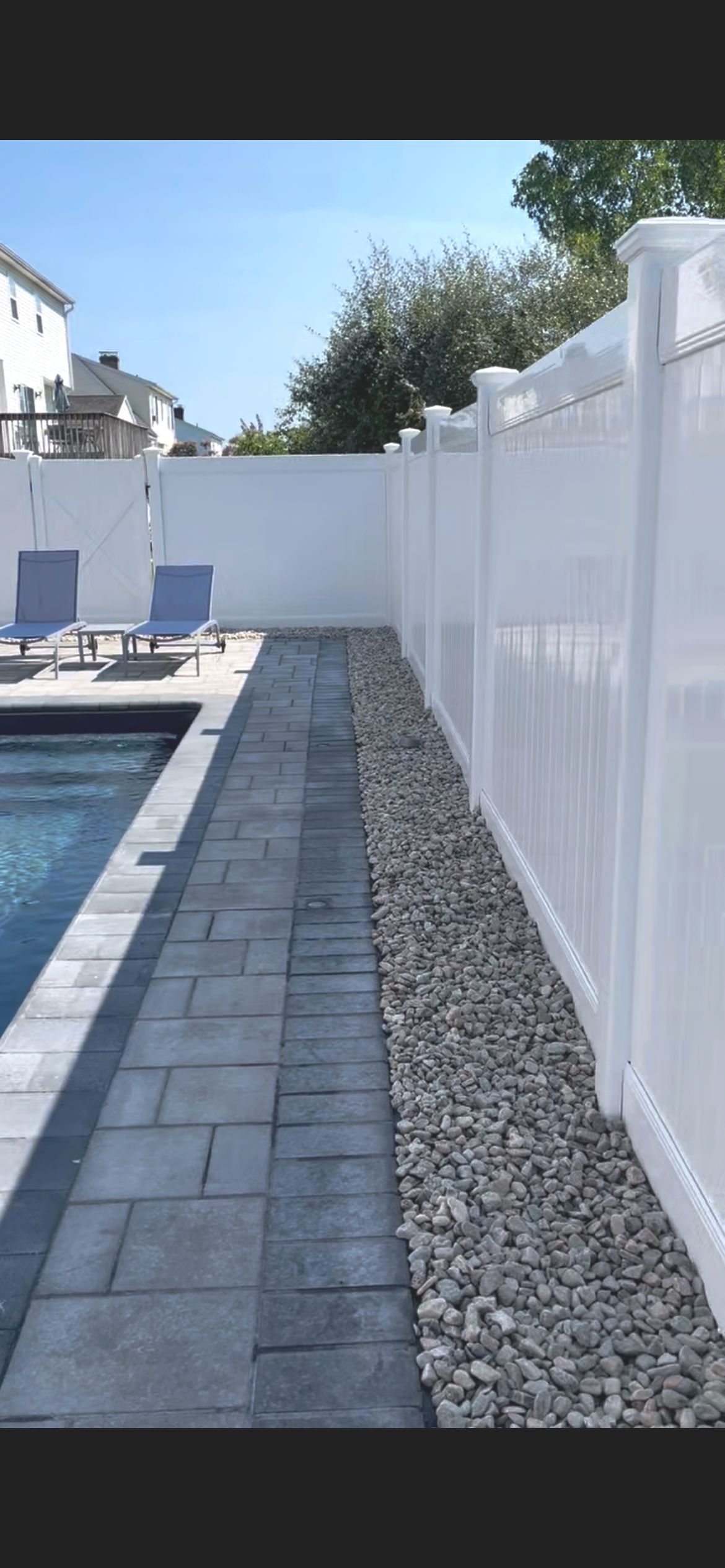 View of a backyard with a swimming pool, two lounge chairs, a paved walkway, a white privacy fence, and a gravel strip beside the fence.