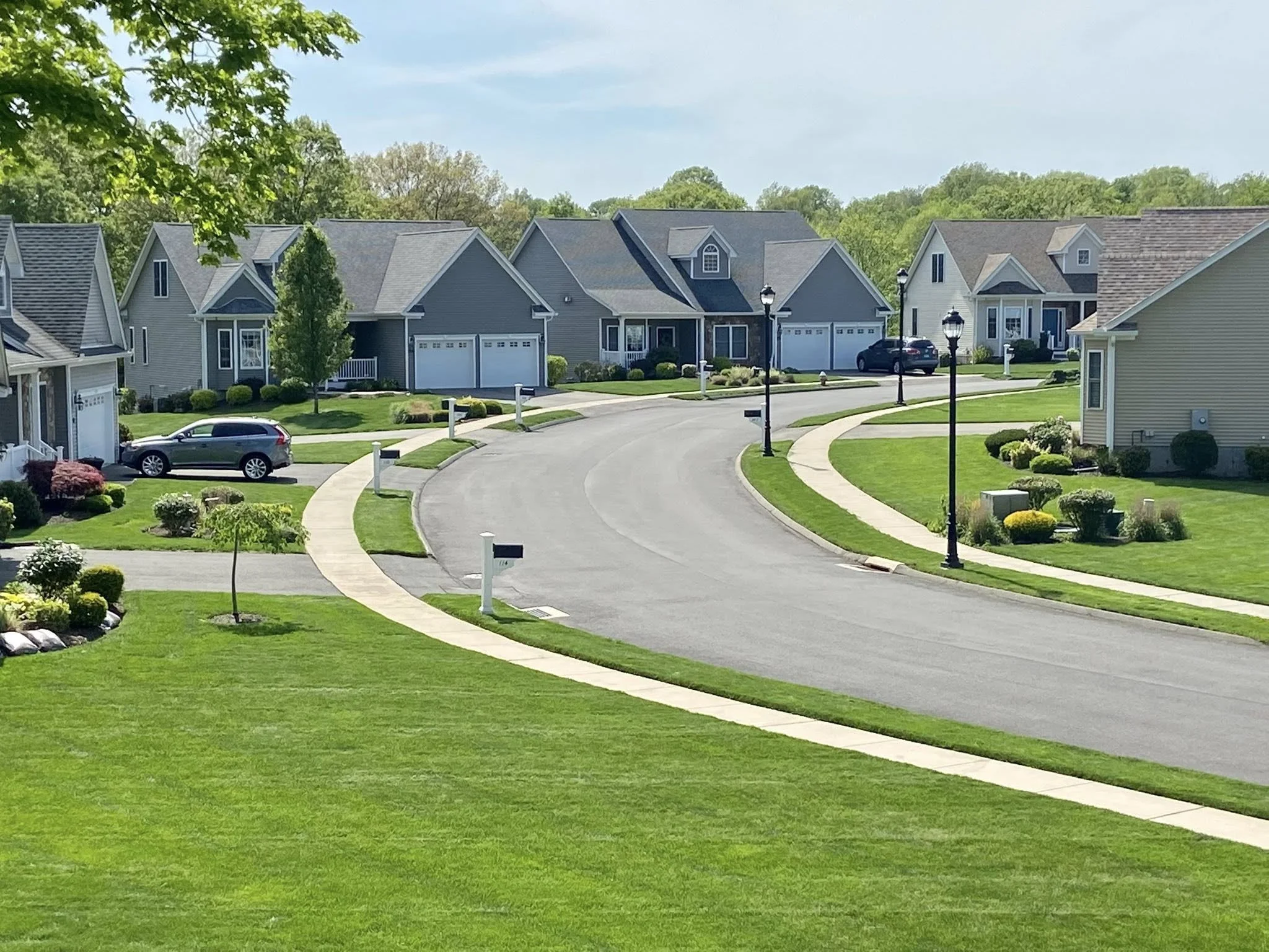 A suburban neighborhood with curved streets, well-manicured lawns, trees, and houses with garages and driveways. A few cars are parked outside.