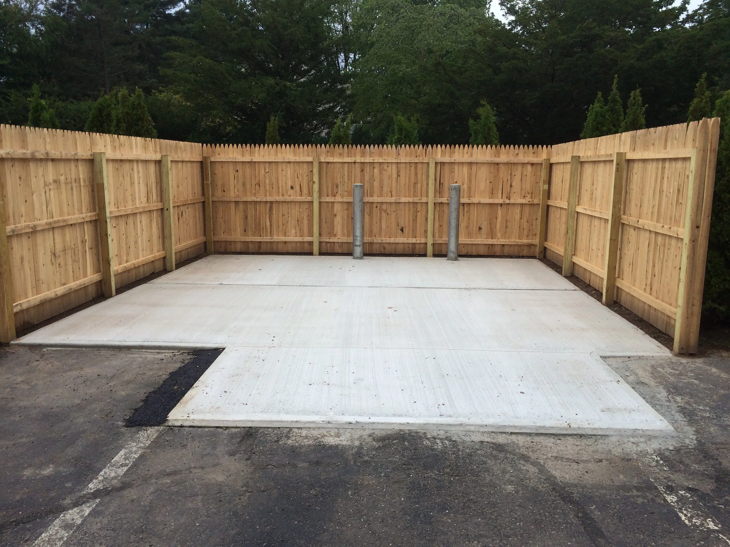 A newly paved concrete parking space with a wooden fence and two metal bollards in the background.