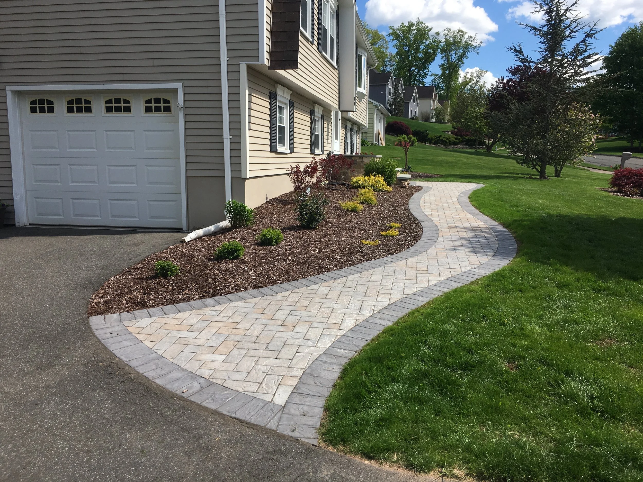 A curved garden bed with newly planted bushes and small plants, mulched with dark wood chips, and a paved walkway opposite a house with beige siding and a white garage door, on a sunny day with a blue sky and green trees in the background.