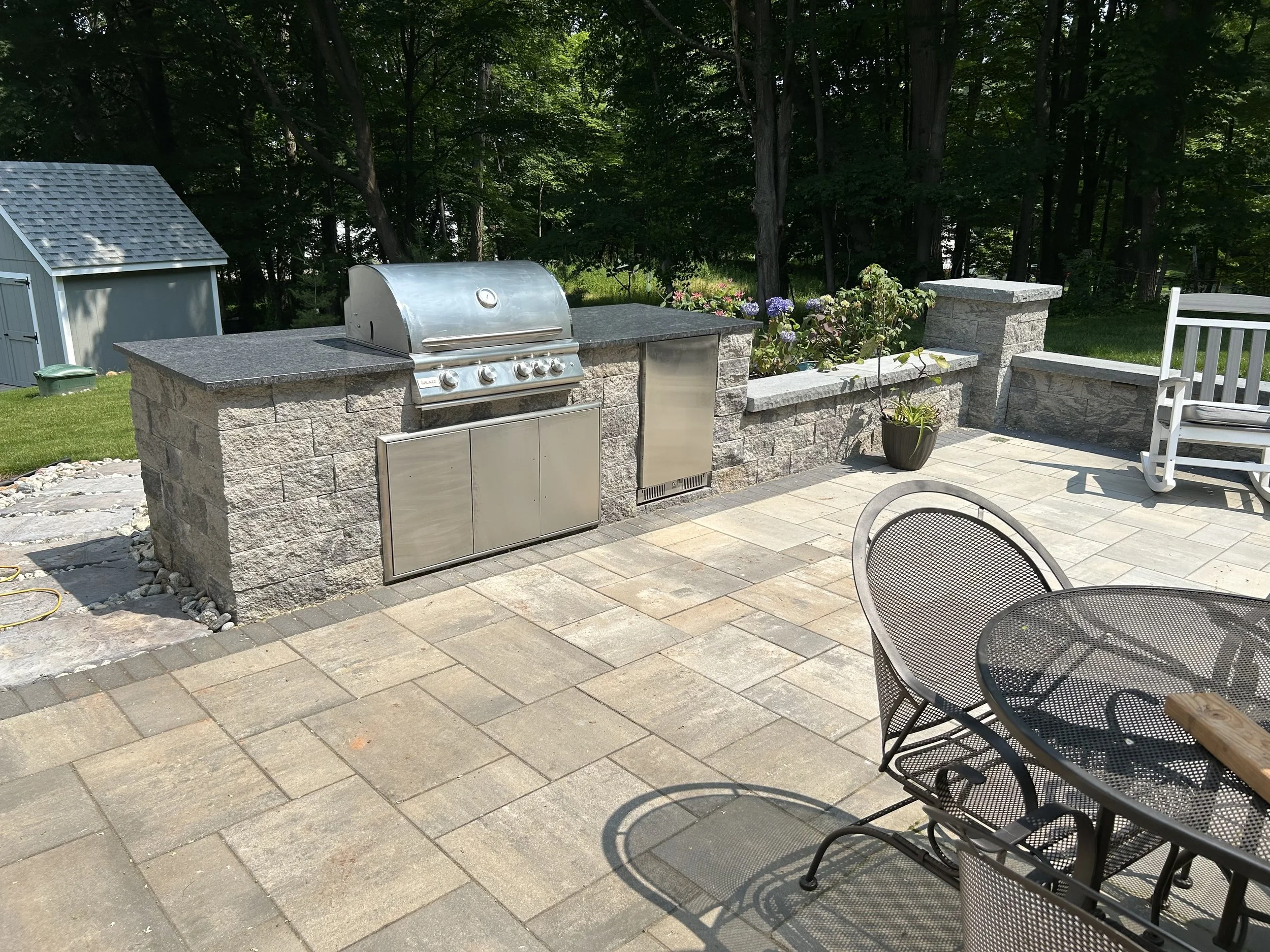 Outdoor patio with built-in stone countertop and grill, potted plants, a white rocking chair, and metal patio table and chairs, with a grassy yard and trees in the background.