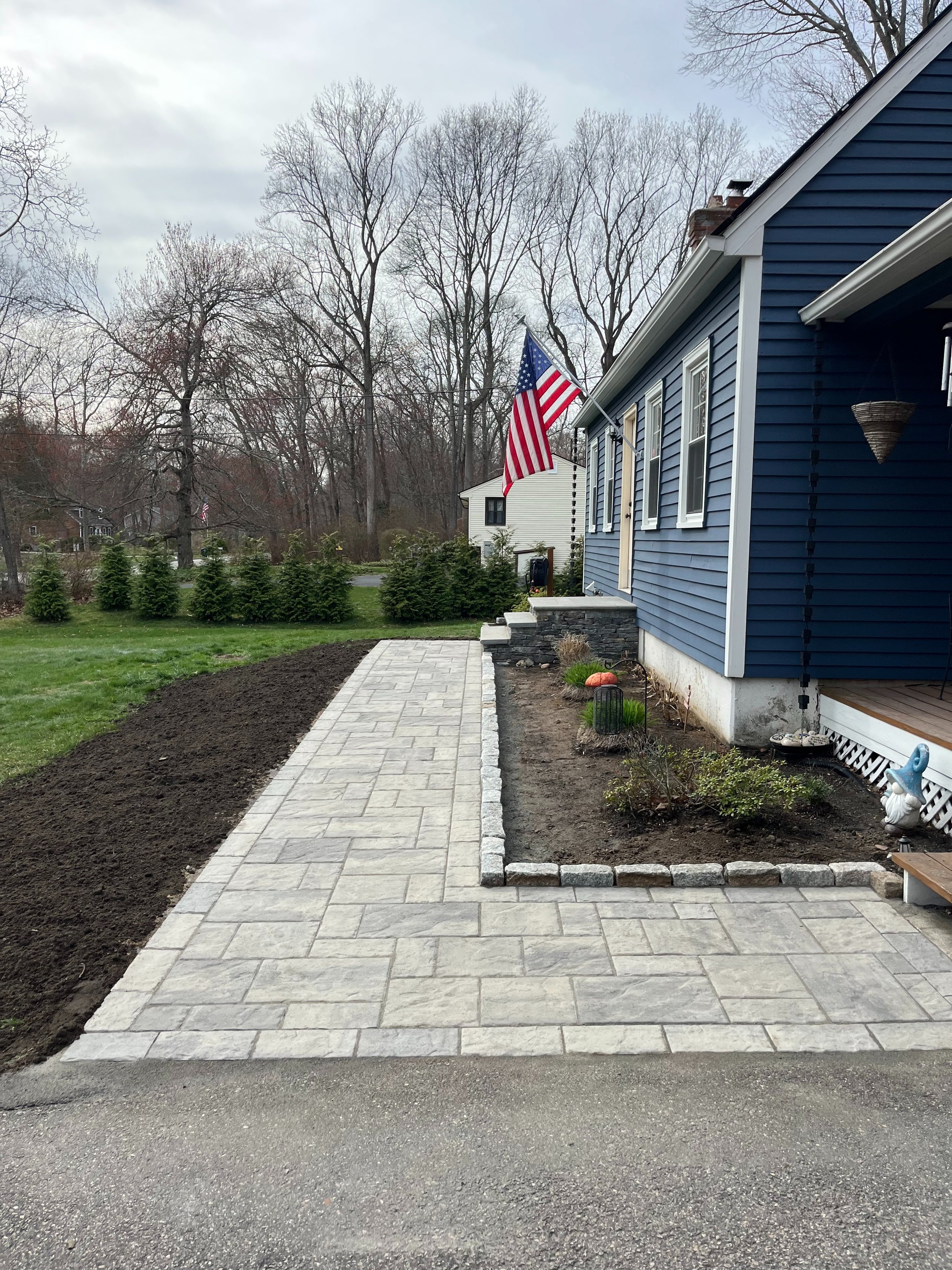 A newly paved stone walkway leading to a blue house with an American flag, surrounded by a garden bed with small plants and pumpkins, and a row of small evergreen trees in the background.