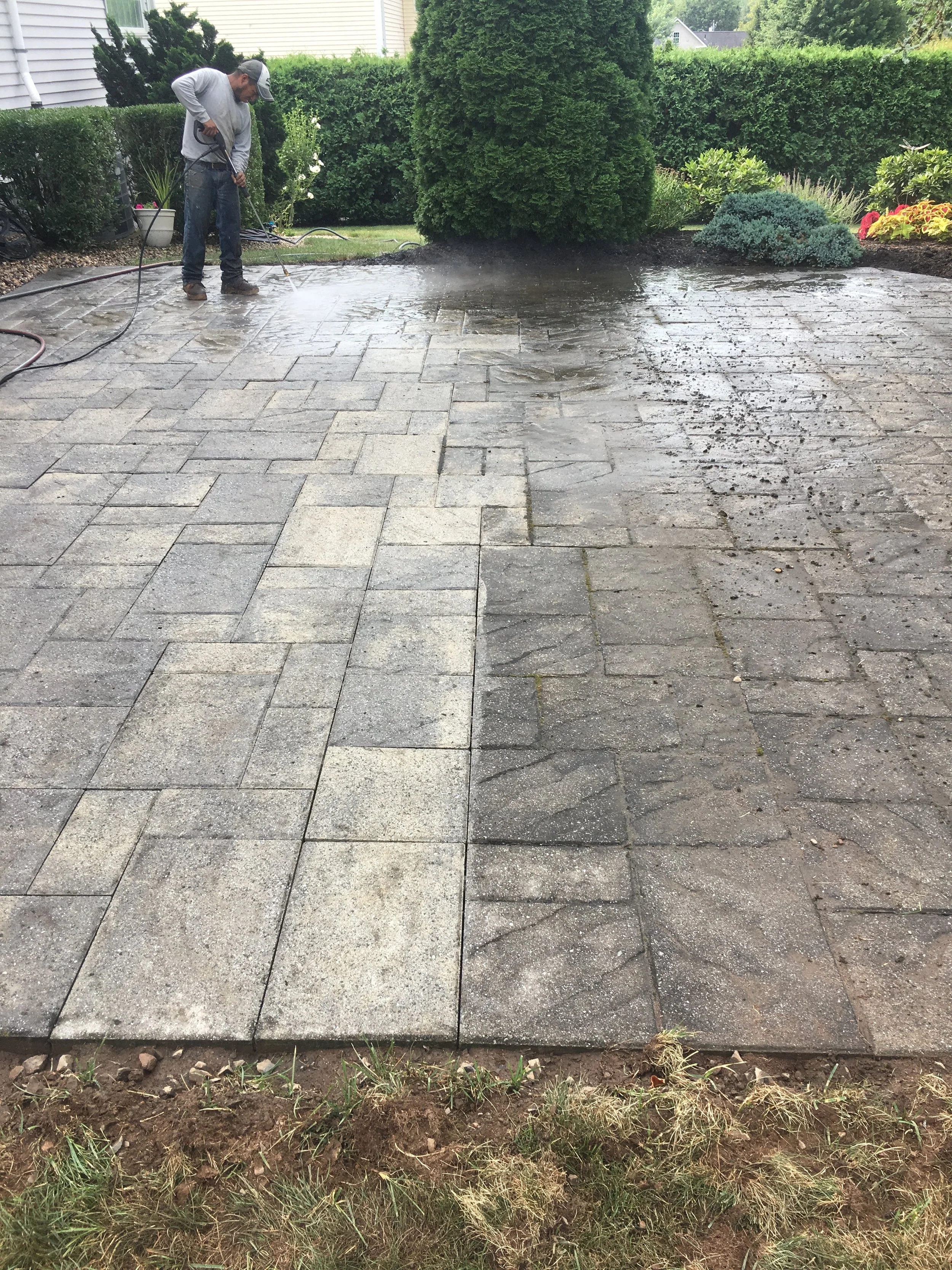 A man power washing a stone patio in a backyard with green bushes and plants.