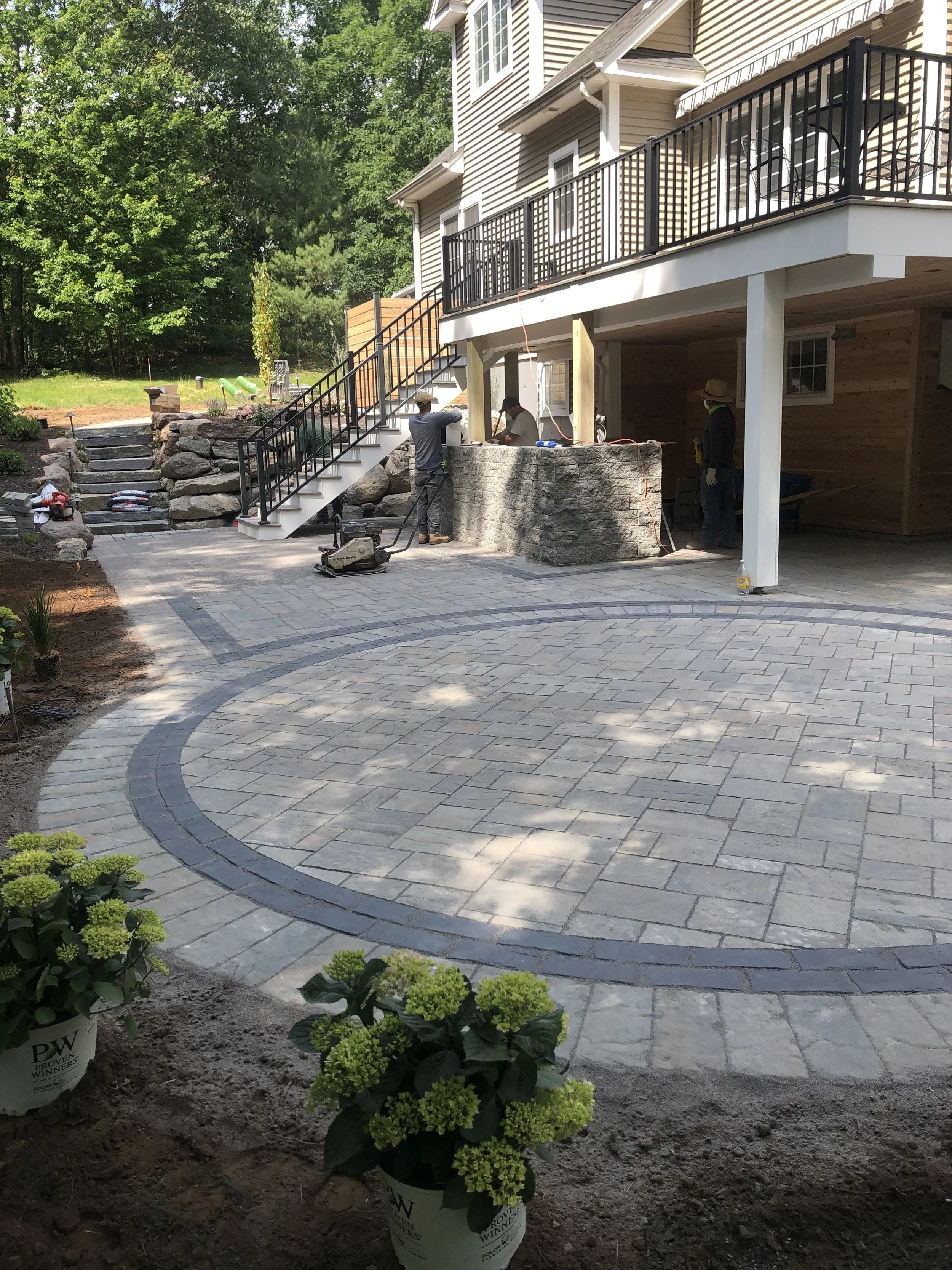 A newly paved outdoor patio with a circular pattern, surrounded by landscaping, and a construction crew working on a stone wall beneath a house with a deck.