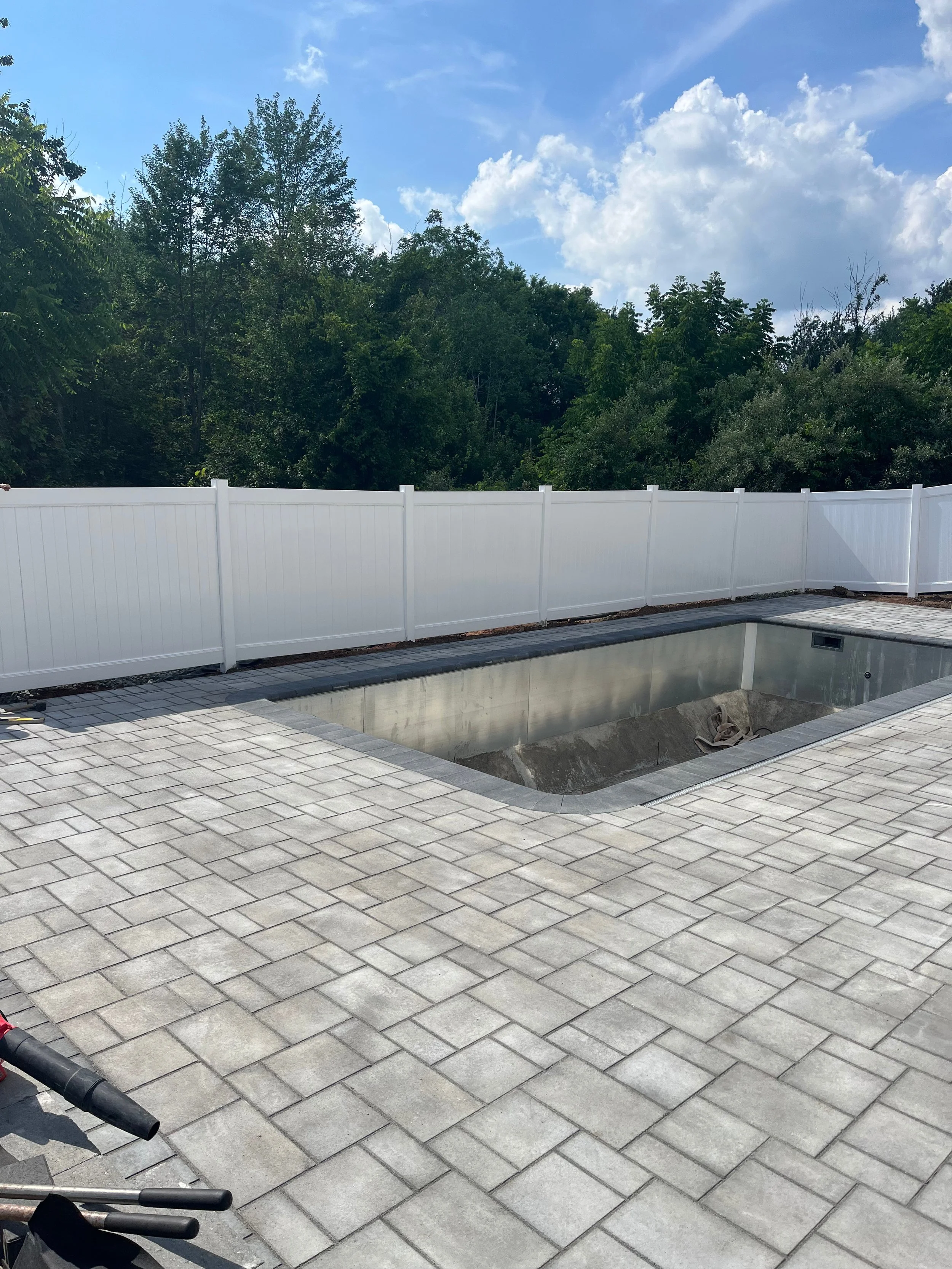 A backyard with a in-ground pool under construction, surrounded by grey paver patio and a white privacy fence