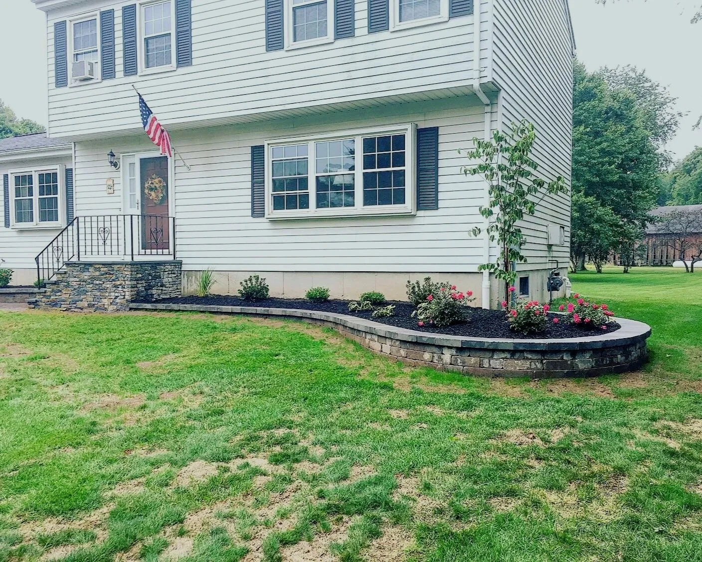 A white two-story house with black shutters, an American flag, and a small front garden with flowers and a young tree. There is a porch with stone steps and a black railing.