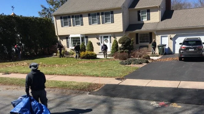 A suburban house with several people standing and walking in the front yard and driveway. One person is standing near the sidewalk holding a blue bag or tarp. The house has beige siding, multiple large windows, and a white garage door. The yard has bushes and trees, with a clear blue sky overhead.