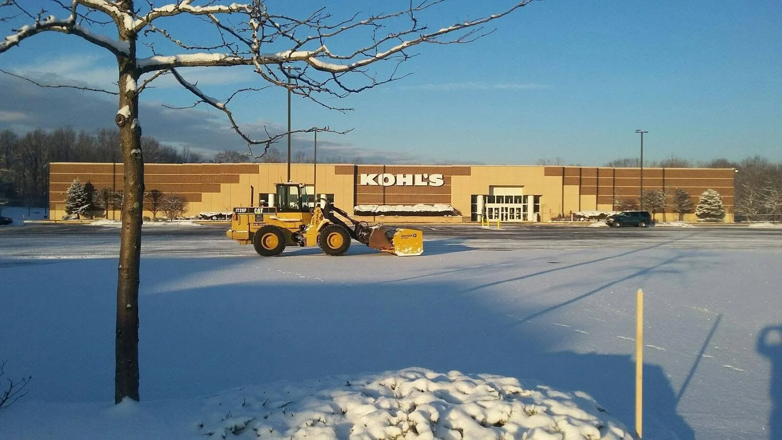 A snow-covered parking lot in front of a Kohl's store with a construction loader clearing snow. The store is beige with large Kohl's signage and a row of trees and parked cars visible in the background, under a partly cloudy sky.