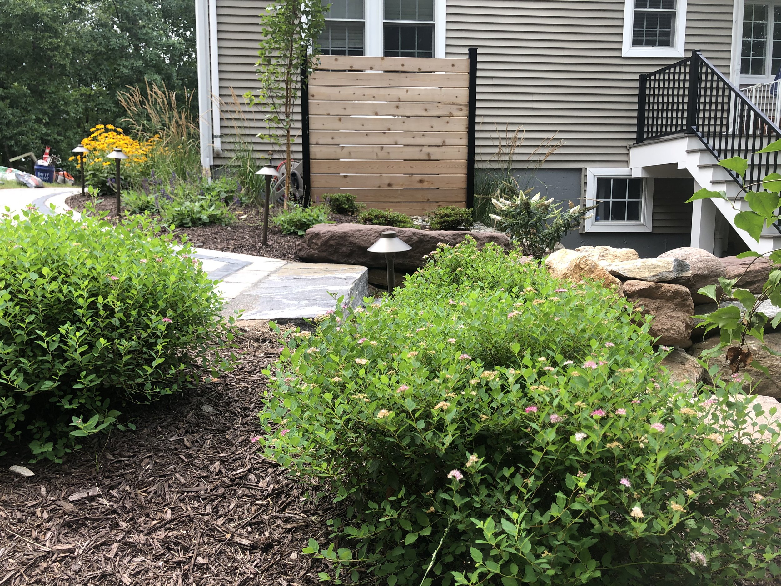 A landscaped backyard garden with green bushes, flowering plants, rock borders, a stone pathway, and a wooden privacy screen.