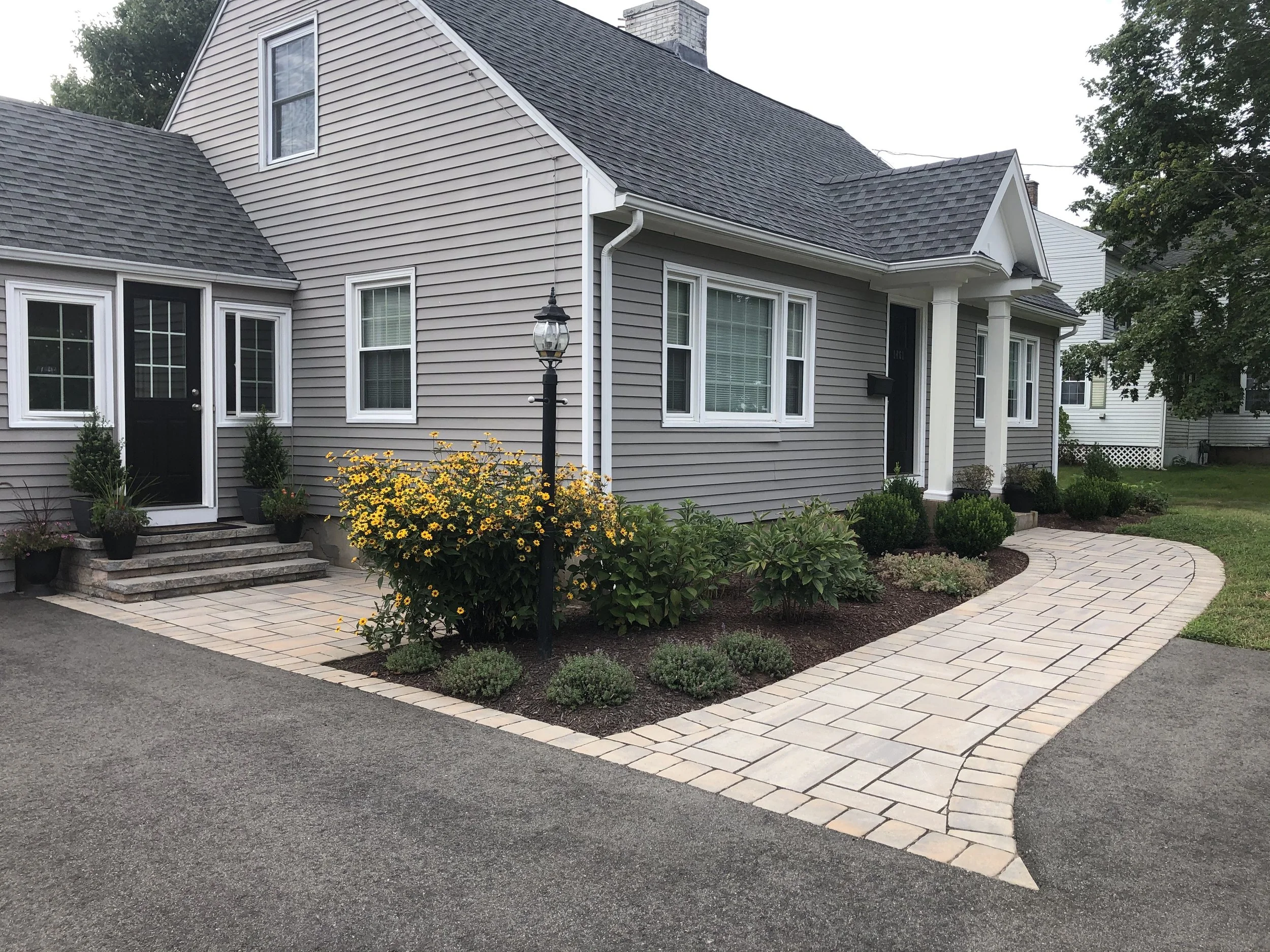 Front yard of a gray house with a curved stone walkway, black door, and landscaped garden with yellow flowers and shrubbery.